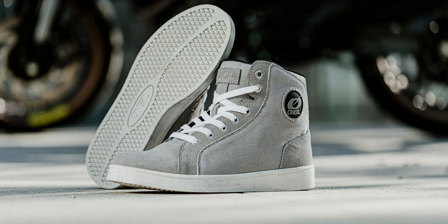 Ein Paar graue High-Top-Sneaker wird ausgestellt, wobei ein Schuh aufrecht steht und der andere auf der Seite liegt, um die strukturierte Sohle und die Schnürsenkel zu zeigen. Der Hintergrund ist leicht unscharf, um den Fokus auf die Sneaker zu richten.