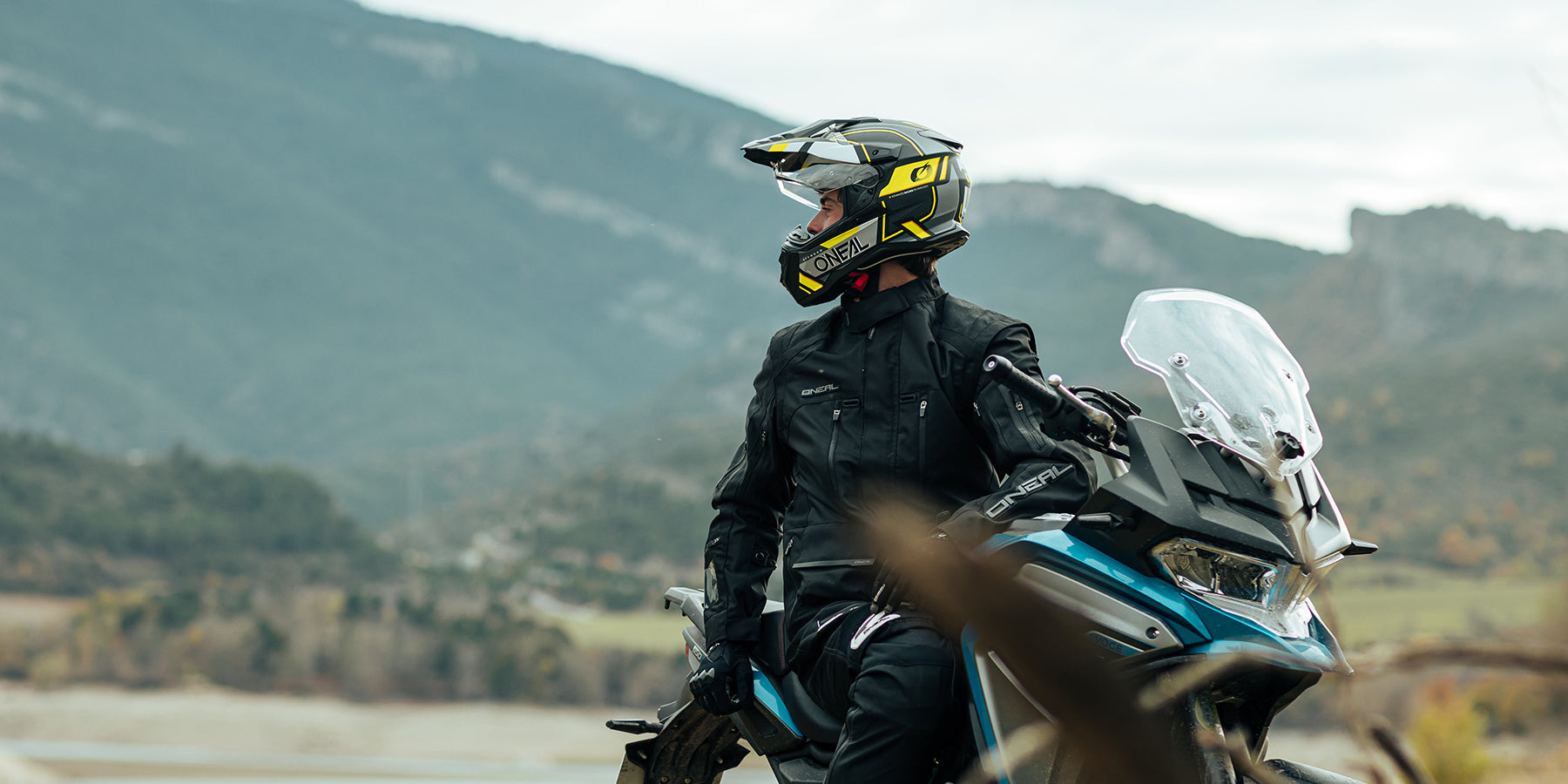 Eine Person in voller Motorradmontur, einschließlich einer schwarzen Jacke und einem Helm mit gelben Akzenten, steht neben einem geparkten Adventure-Motorrad. Sie stehen vor einer Berglandschaft, mit einem sichtbaren Tal und etwas Laub im Hintergrund.