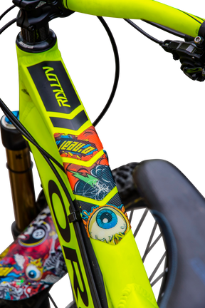 Primo piano di un set di FRAME GUARDS di AZONIC con adesivi colorati in stile fumetto, come ad esempio un vistoso bulbo oculare blu e una grafica da supereroe, che adornano il telaio giallo di una bicicletta con il manubrio nero e la forcella della sospensione anteriore.