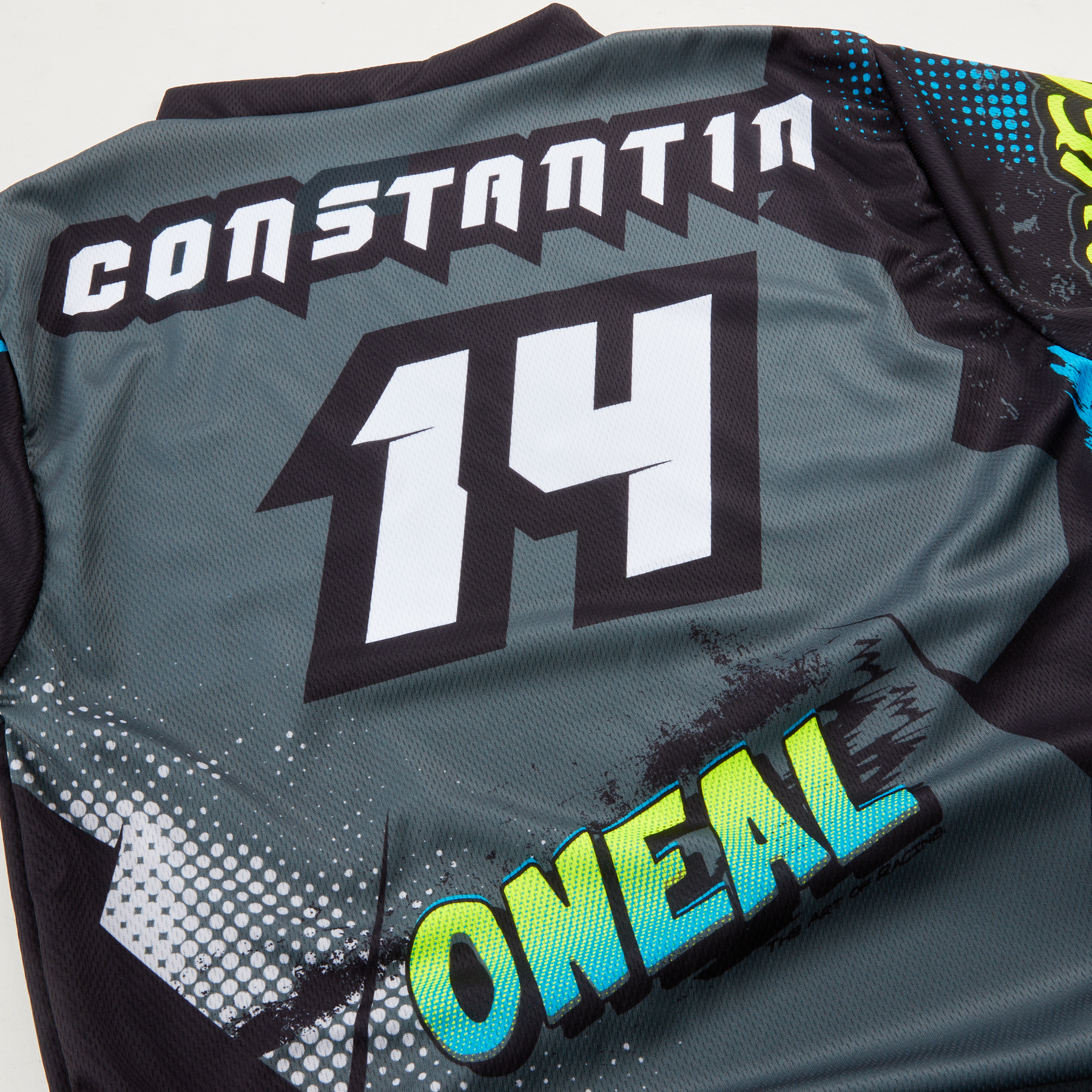 Eine Nahaufnahme eines O'NEAL Custom Trikots Kinder VILLAIN mit "CONSTANT1N" und der Nummer "14" in fettgedrucktem Weiß, sowie "ONEAL" in leuchtendem Grün und Blau auf dem unteren Teil dieses O'NEAL-Trikots.
