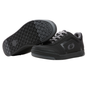 A pair of O'NEAL PINNED FLAT pedal shoes in black with a dark gray sole, featuring a hexagonal grip for flat-pedal riding. One shoe stands upright to show the side and laces; the other tilts to show the textured sole.