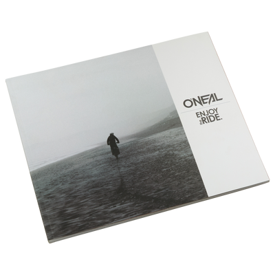 The closed O'NEAL ENJOY THE RIDE BOOK shows a lonely figure on a foggy beach that evokes emotions. The O'NEAL logo and the words "ENJOY THE RIDE." in black on white can be seen on the right-hand side. Brand: O'NEAL. Product: O'NEAL ENJOY THE RIDE BOOK (FREE GOODIE WAGON).