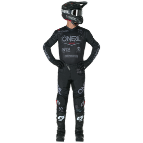 Une personne portant tout l'équipement noir d'O'NEAL, y compris le pantalon O'NEAL ELEMENT Pants BRAND, le casque, les gants et les bottes, se tient debout, les mains sur les hanches, et regarde légèrement vers la gauche.