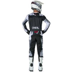 A rider in black, white and gray clothing stands with his hands on his hips and his eyes facing forward. He is wearing a full O'NEAL helmet, boots and the O'NEAL ELEMENT Pants ROLLER with red logo accents against a black background.