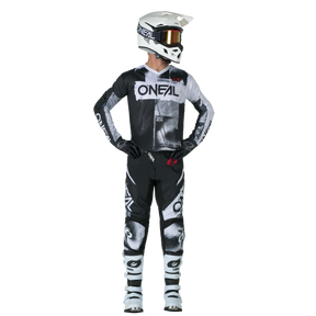 A motocross rider stands upright and looks to the side. He is wearing the O'NEAL ELEMENT Pants ROLLER as well as a matching black and white O'NEAL helmet, goggles, gloves and boots.