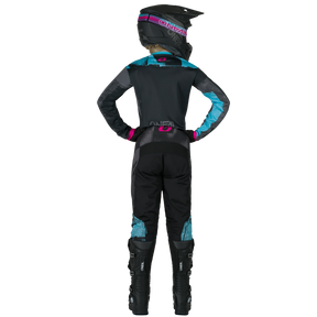 Una persona está de pie con las manos en las caderas y lleva puesto el pantalón de mujer ROLLER O'NEAL Element de O'NEAL, un mono de motocross negro, azul y rosa, un casco y botas de motocross negras.