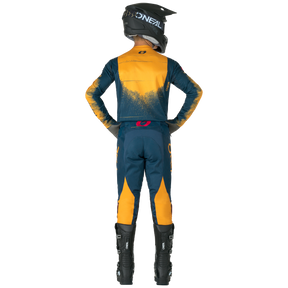 Una persona vestida con PANTALONES O'NEAL Hardwear SWIPE y un traje de motocross O'NEAL a juego, casco y botas negras, de pie, con las manos en las caderas, de espaldas a la cámara.