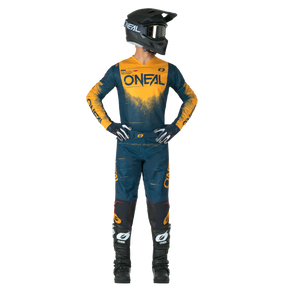 Una persona está de pie con las manos en las caderas y lleva un conjunto completo de motocross con casco negro, camiseta O'NEAL azul y naranja, guantes y botas negras y pantalones O'NEAL Hardwear SWIPE.