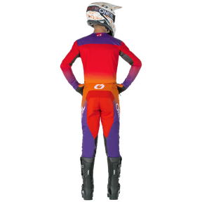 Une personne vêtue d'une combinaison de motocross O'NEAL MAYHEM pantalon HEXX rouge, orange et violet avec des bottes noires et un casque blanc se tient debout, les mains sur les hanches. La tenue présente le branding O'NEAL sur le casque, le dos et les jambes.