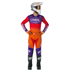 Un coureur de motocross avec un casque blanc, un maillot O'NEAL rouge et orange, un pantalon O'NEAL MAYHEM HEXX avec des accents violets et des bottes noires se tient debout, les mains sur les hanches, et regarde de côté.