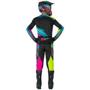 Eine Person, die die O'NEAL MAYHEM Pants RESEDA von O'NEAL mit leuchtenden blauen, pinken, gelben und schwarzen Mustern trägt, steht mit den Händen auf den Hüften von der Kamera abgewandt in einem kompletten Motocross-Outfit.