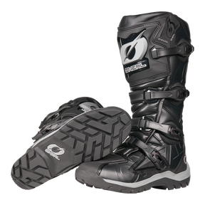 La bota O'NEAL RMX Adventure en negro y gris tiene suelas robustas, hebillas protectoras y espinilleras reforzadas. Una bota se mantiene erguida, mientras que la otra se coloca de lado para resaltar el perfil: ideal para los entusiastas de los deportes de motor.