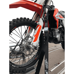 Gros plan sur une moto tout-terrain rouge et blanche attachée à une remorque avec des sangles d'arrimage O'NEAL DE LUXE LOGO sur la fourche avant. Les sangles de la marque O'NEAL maintiennent le vélo, la roue avant et la suspension étant clairement visibles.