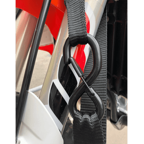 Gros plan sur un harnais O'NEAL DE LUXE LOGO en nylon noir avec un crochet en S en plastique fixé à la fourche avant d'une moto, avec des soft hook tie downs et des détails rouges, noirs et blancs en arrière-plan.