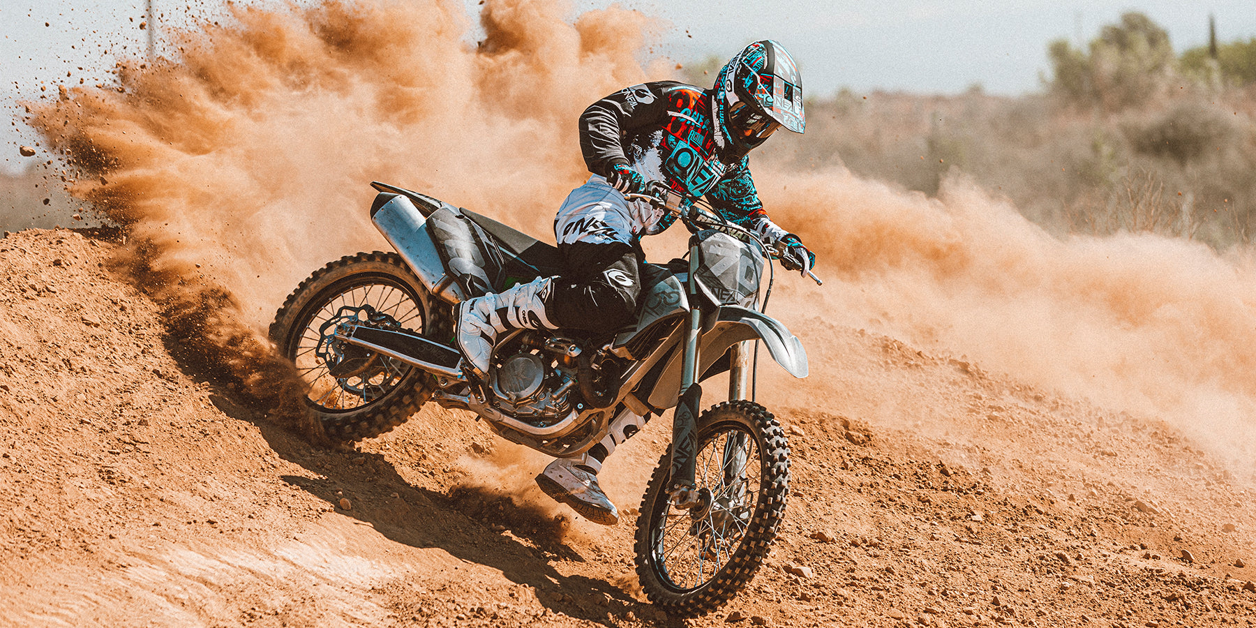 Ein Motocrossfahrer in voller Montur driftet durch eine staubige Kurve auf einer unbefestigten Strecke. Der Fahrer lehnt sich in die Kurve und wirbelt eine Staubwolke hinter dem Motorrad auf, was die Intensität und Geschwindigkeit des Sports veranschaulicht. Trockenes Gelände und ein unscharfer Hintergrund deuten auf viel Bewegung hin.