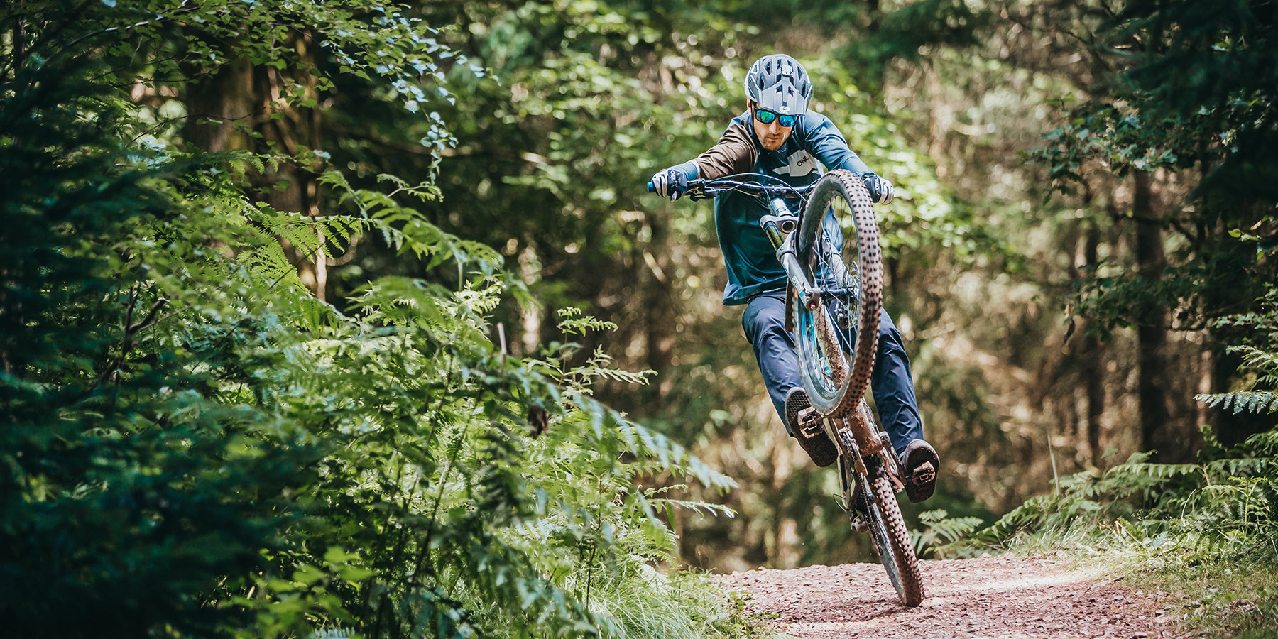 Ein Radfahrer mit Helm und Sonnenbrille macht einen Wheelie, während er mit seinem Mountainbike auf einem Feldweg durch einen dichten, grünen Wald fährt. Der Fahrer ist in Bewegung, balanciert auf dem Hinterrad und ist von üppiger Vegetation umgeben.
