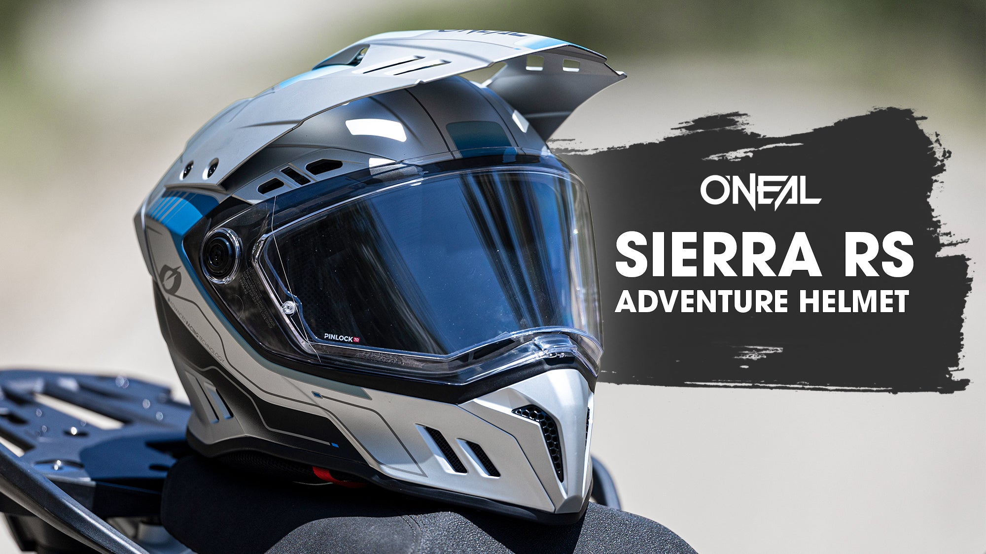 Ein silberner und schwarzer O'Neal Sierra RS Abenteuerhelm sitzt auf einem Motorradsitz, daneben steht der Text "ONEAL SIERRA RS ADVENTURE HELMET" auf einem schwarzen Farbstrich-Hintergrund.