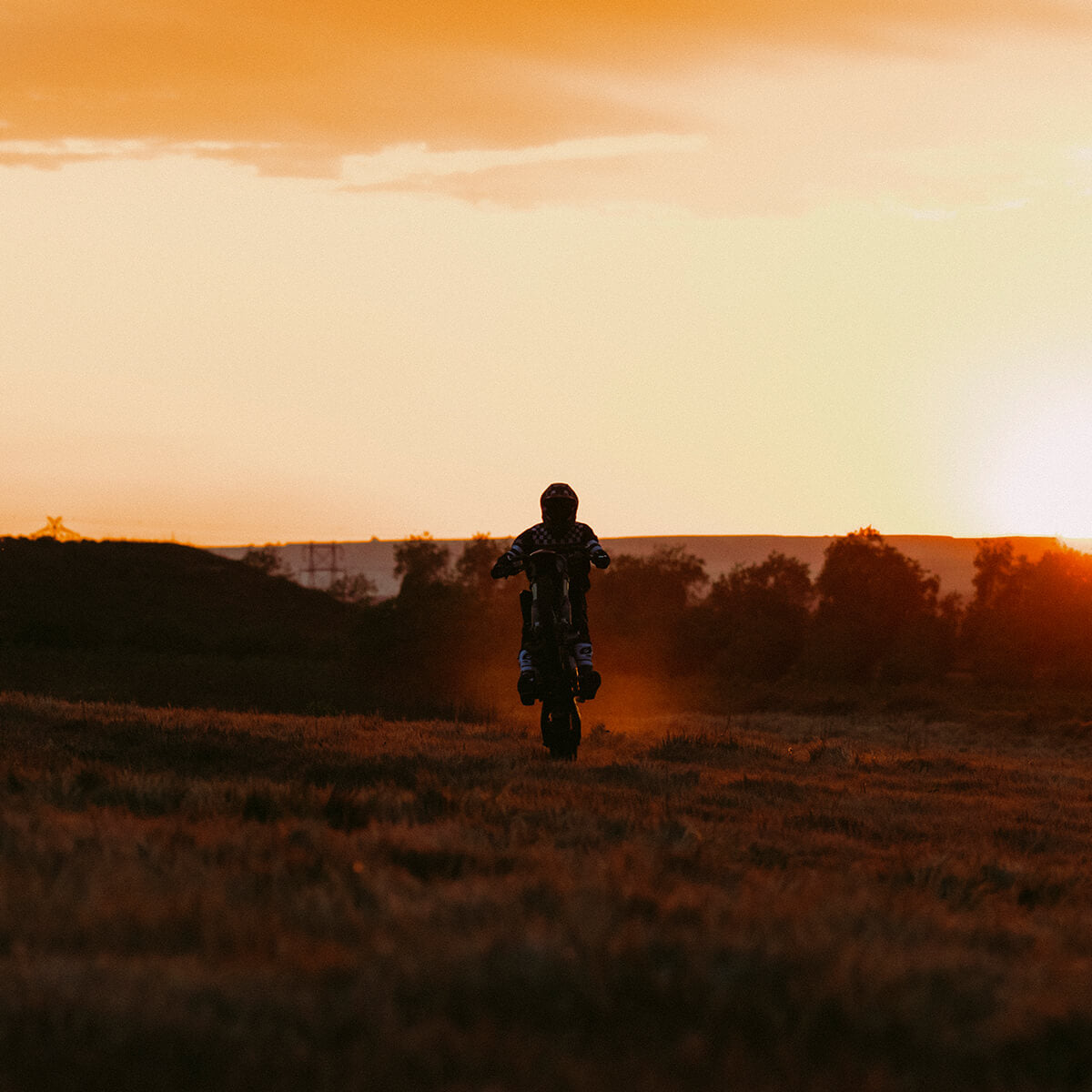 Eine Person macht bei Sonnenuntergang mit einem Motorrad einen Wheelie über eine Wiese. Der Himmel hat einen warmen Orangeton und die Silhouette des Fahrers hebt sich deutlich vom leuchtenden Horizont ab. Im Hintergrund sind schwach Bäume und Hügel zu erkennen.
