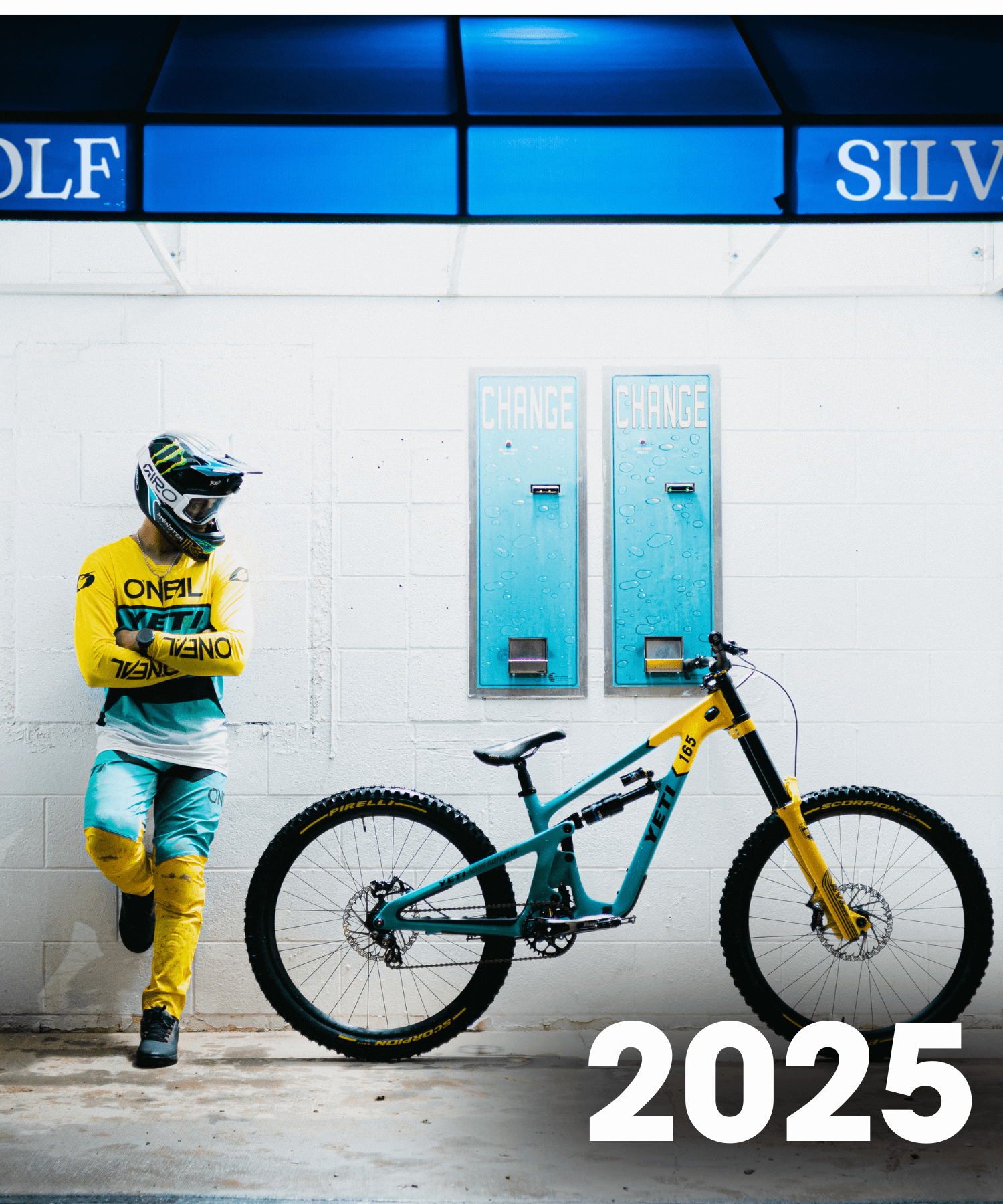 Ein Radfahrer in gelber und türkisfarbener Kleidung lehnt an einer weißen Wand neben einem passenden Mountainbike, über dem zwei Wechselautomaten und eine blaue Markise angebracht sind. Die Zahl "2025" ist in fetter weißer Schrift unten rechts zu lesen.