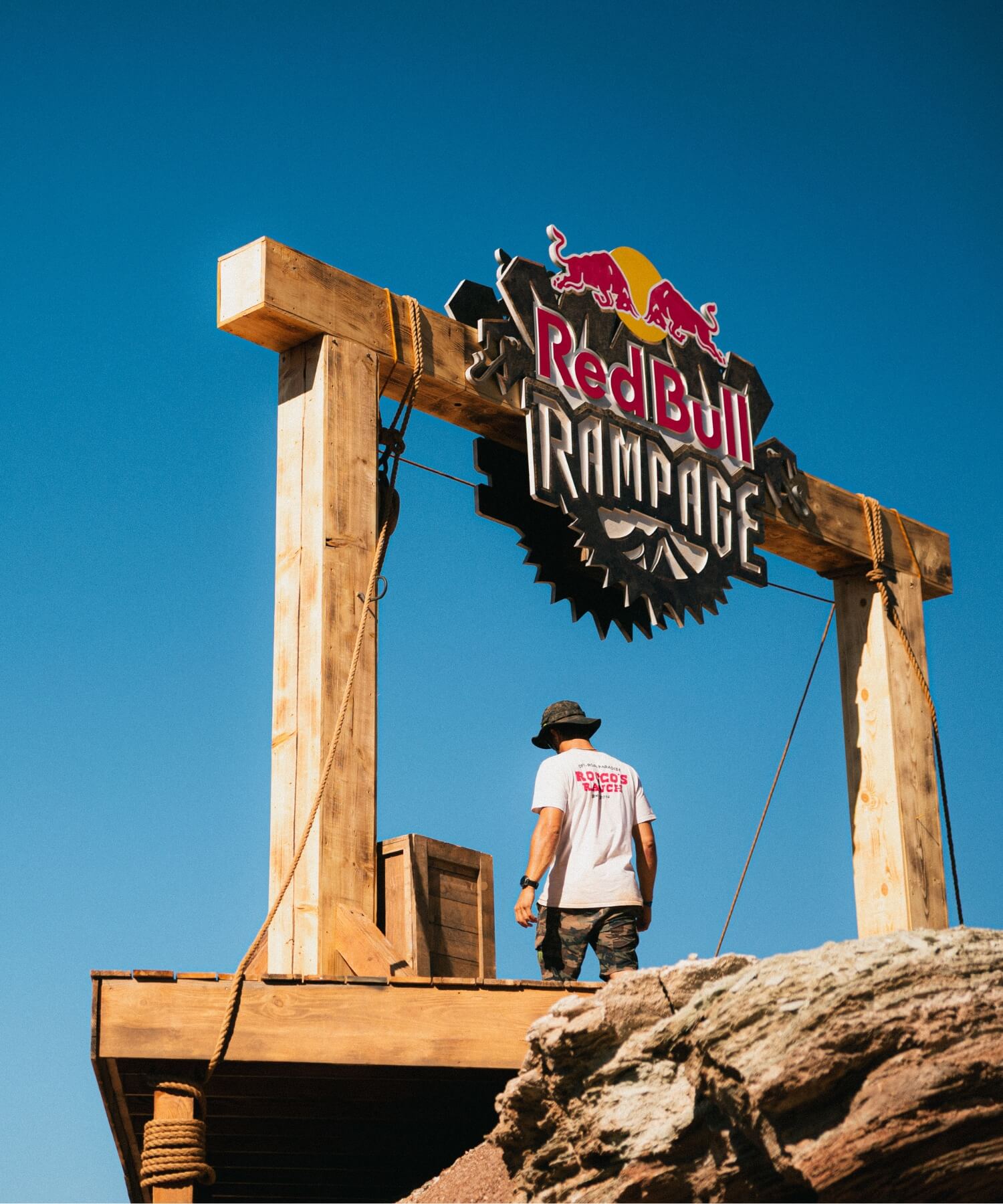 Eine Person in einem weißen T-Shirt steht auf einem Holzpodest unter einem großen Red Bull Rampage"-Schild vor einem strahlend blauen Himmel, mit felsigem Gelände im Vordergrund.