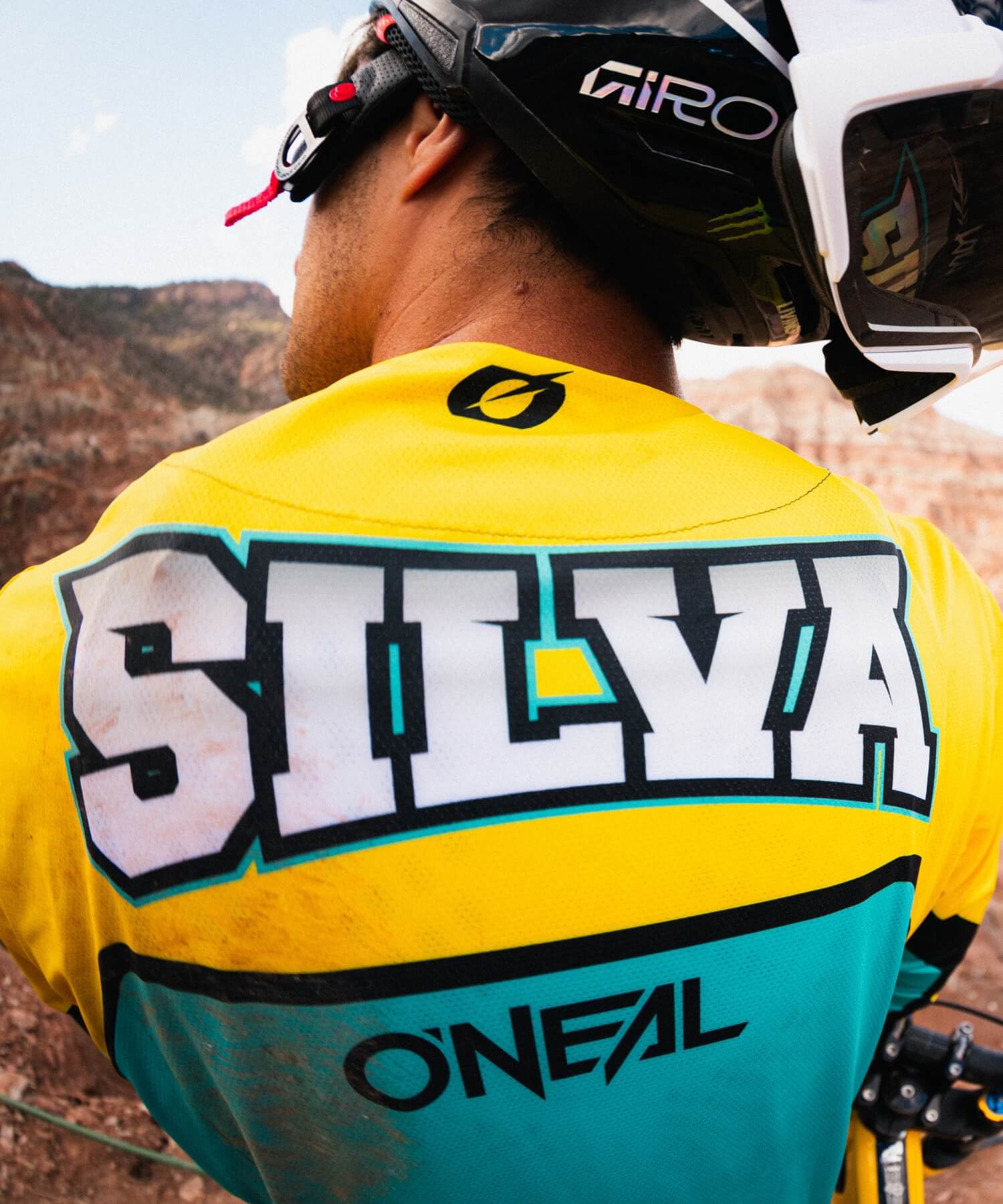 Ein Mountainbiker, der ein gelbes und blaugrünes Trikot mit dem Namen "SILVA" und der Marke "O'NEAL" auf dem Rücken sowie einen schwarzen Giro-Helm trägt, steht im Freien vor einem felsigen Gelände im Hintergrund.