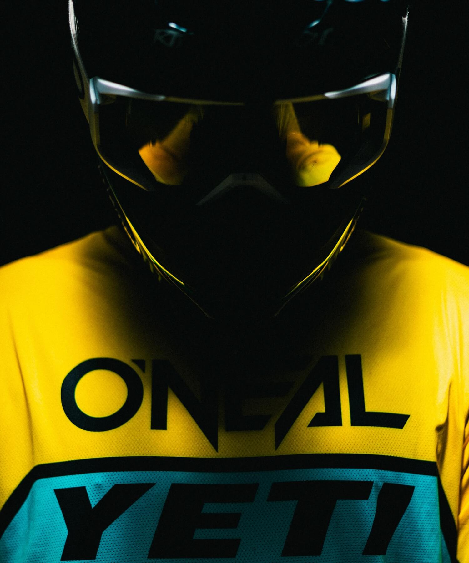 Eine Person, die einen schwarzen Helm mit Visier und ein gelbes Trikot mit den Logos "O'NEAL" und "YETI" trägt, steht vor einem dunklen, teilweise von oben beleuchteten Hintergrund.