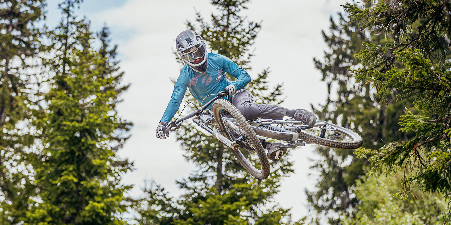 Ein Radfahrer in einem blauen Langarmshirt, grauen Hosen und einem Helm vollführt vor einer Kulisse aus hohen, grünen Bäumen einen Hochsprung auf einem Mountainbike. Das Fahrrad des Fahrers ist zur Seite geneigt, was die Bewegung und das Können betont, die für den Stunt erforderlich sind.