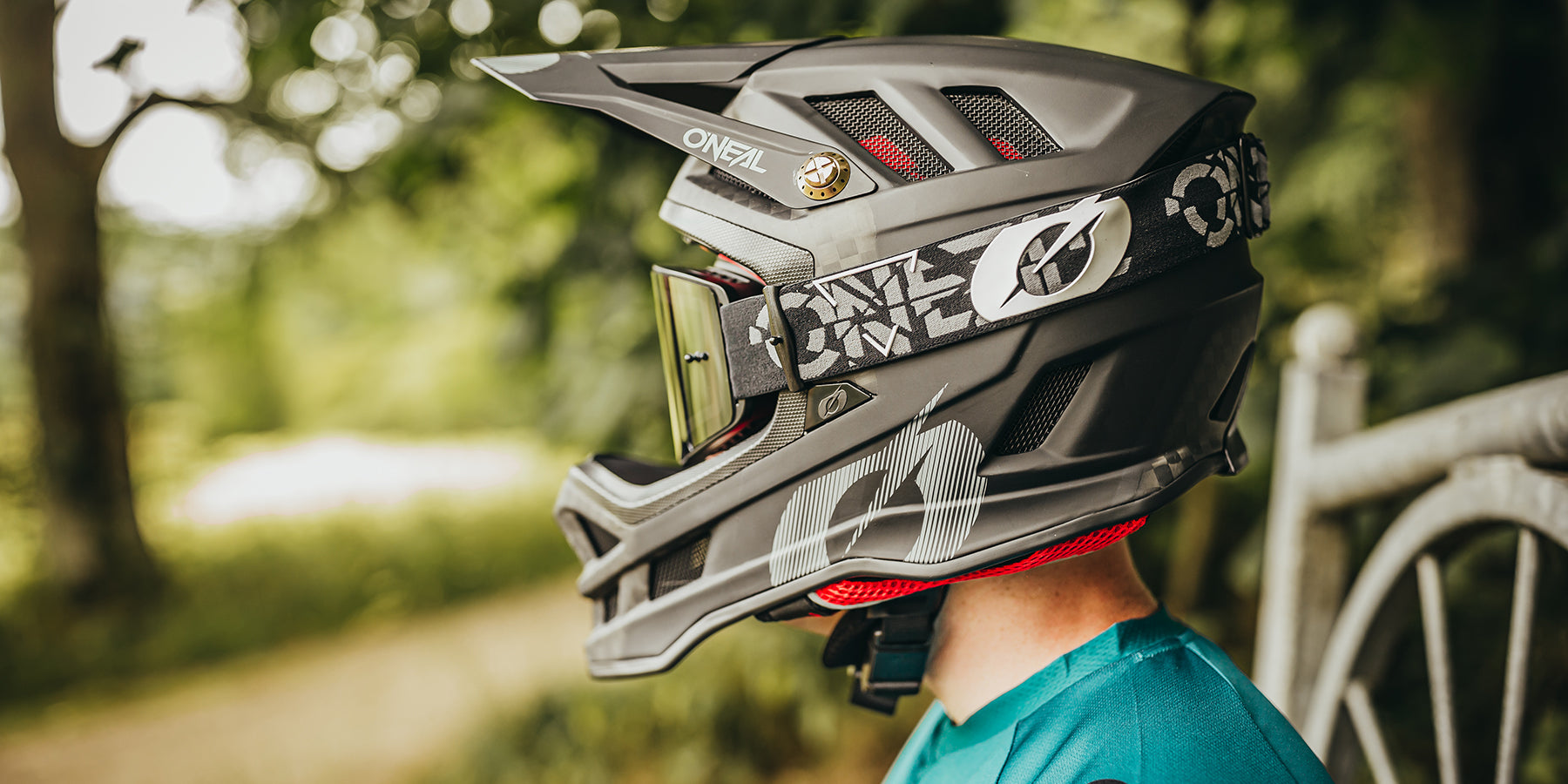 Seitenansicht einer Person, die einen schwarz-grauen O'Neal-Integralhelm mit Schutzbrille trägt. Der Helm hat ein Visier und ein aufwendiges Design. Die Person trägt ein blaugrünes Hemd und steht im Freien mit verschwommenem Grün im Hintergrund.