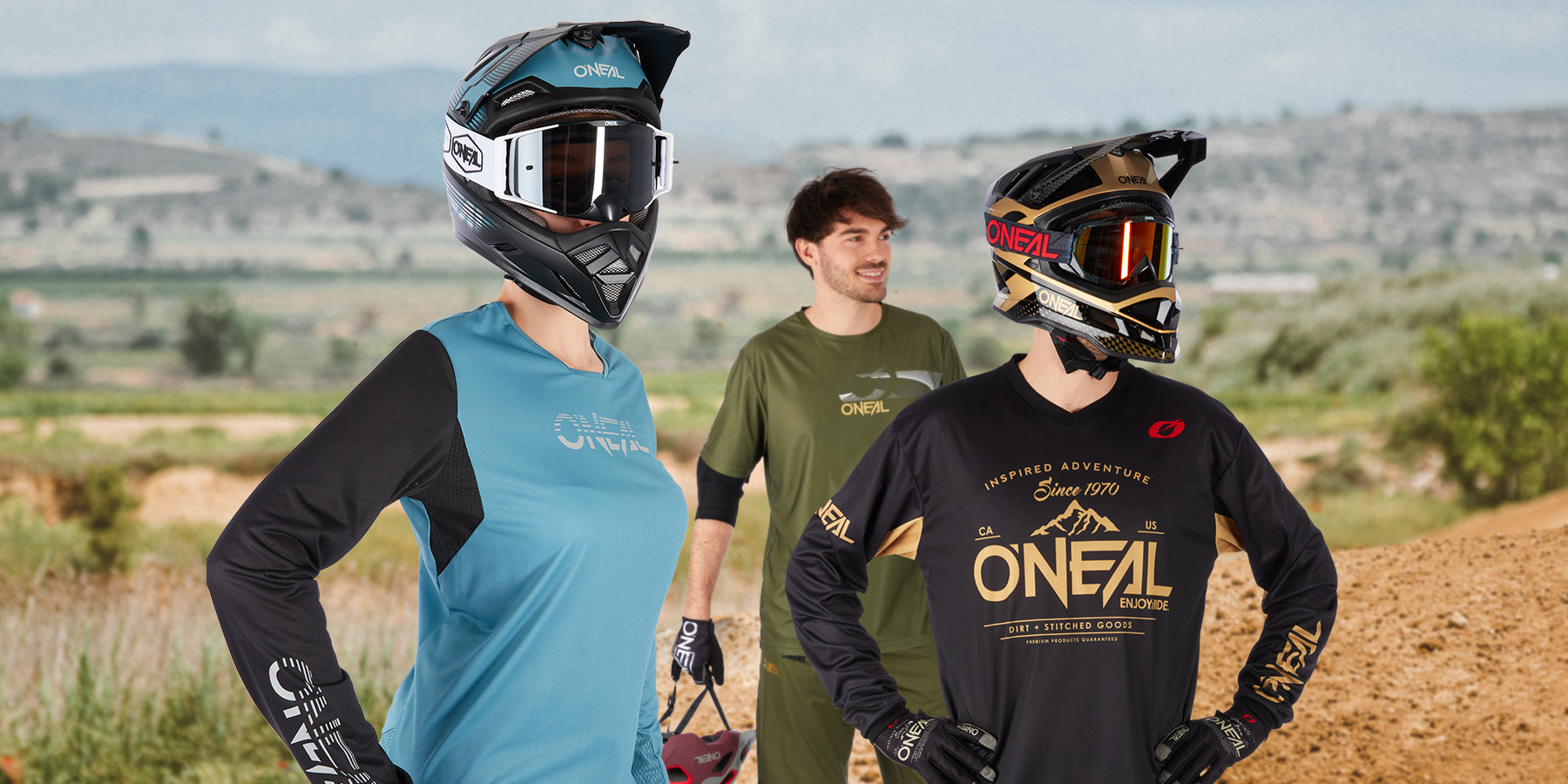 Drei Personen in Motocross-Ausrüstung stehen im Freien. Die beiden im Vordergrund tragen Helme und Trikots (einer in Blau und Schwarz, der andere in Schwarz und Gold), während die Person im Hintergrund, die keinen Helm trägt, ein grünes Trikot trägt und einen Helm hält.