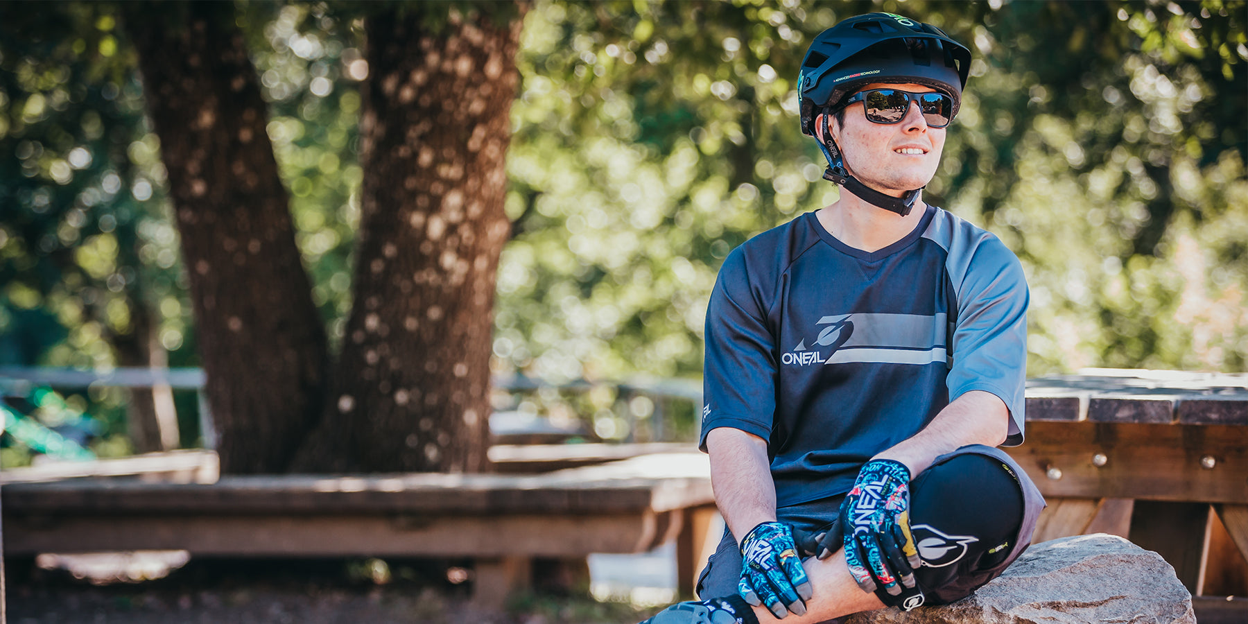 Eine Person in blauem Radsport-Outfit, Handschuhen, Helm und Sonnenbrille sitzt draußen auf einem Felsen. Sie wirkt entspannt, hat ein Bein über das andere geschlagen und blickt in die Ferne. Im Hintergrund sind Bäume und Holzbänke zu sehen, was an eine Park- oder Waldlandschaft erinnert.
