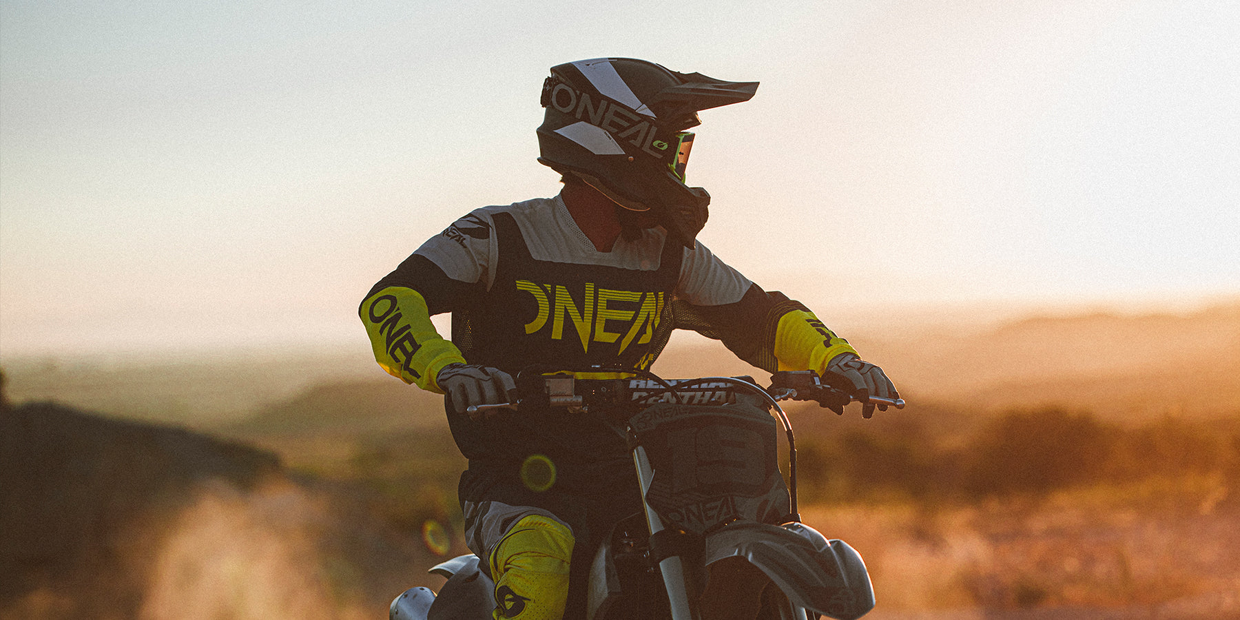 Eine Person in gelb-schwarzer Motocross-Ausrüstung fährt ein Dirtbike in einer sonnigen Umgebung im Freien. Der Fahrer trägt einen Helm mit Visier, Handschuhe und ein Trikot mit der Aufschrift „ONEAL“. Im Hintergrund ist eine dunstige Landschaft mit einem warmen, goldenen Himmel zu sehen.