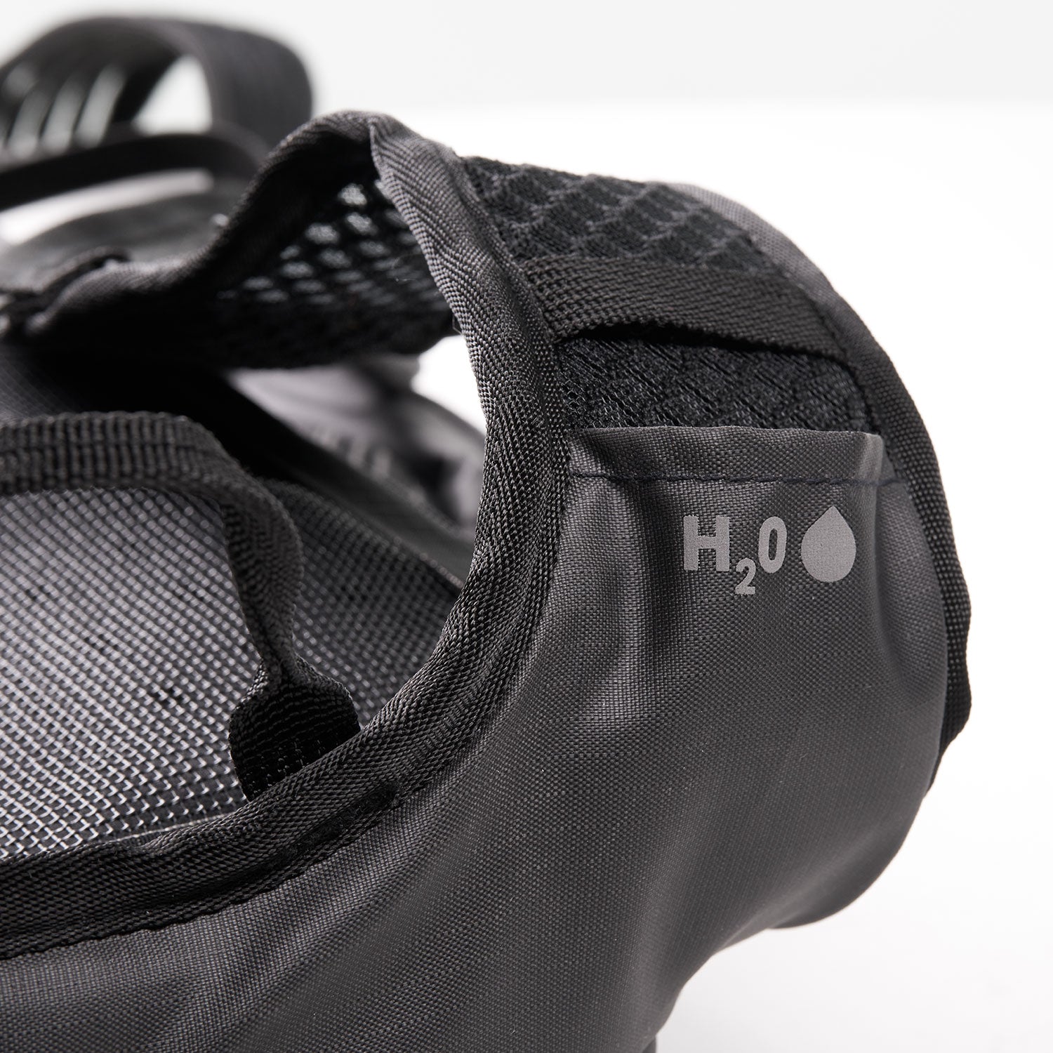 Nahaufnahme eines schwarzen Rucksacks oder einer Weste mit Mesh- und Stoffdetails, die eine kleine Tasche mit der Aufschrift "H2O" und einem Wassertropfensymbol aufweisen, was auf ein Fach für einen Trinkspritzbehälter hinweist.