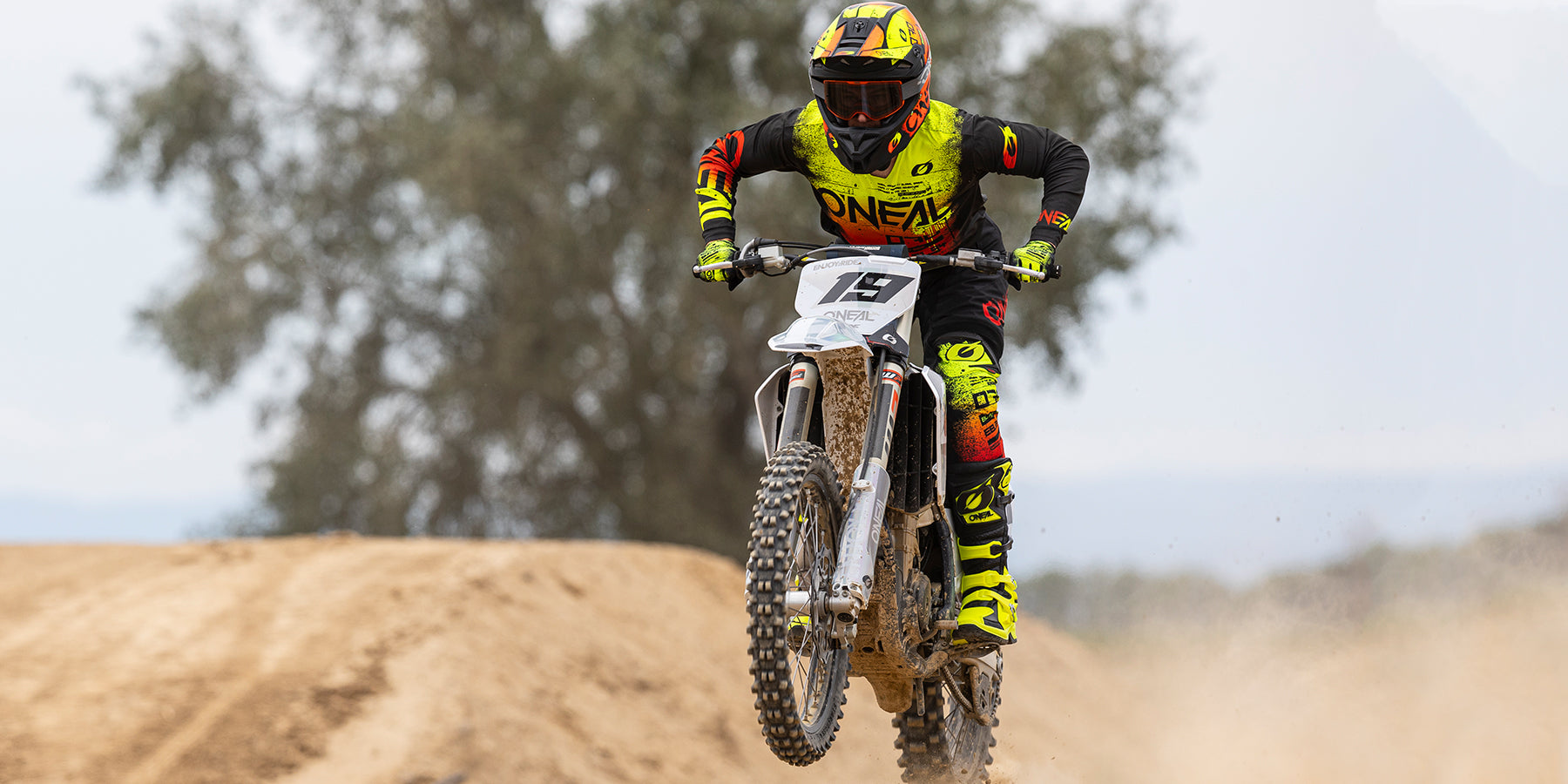 Ein Motocrossfahrer mit gelb-schwarzem Helm und passender Ausrüstung (Nummer 19) ist auf einem Dirtbike in der Luft. Im Hintergrund sind eine sandige Motocross-Strecke und verschwommene Bäume zu sehen.