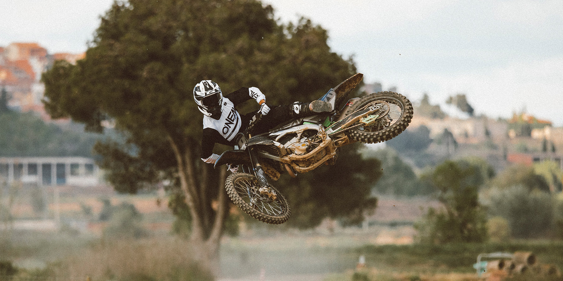 Ein Motocrossfahrer in der Luft, der einen Trick auf einem Dirtbike vorführt. Er trägt Schutzausrüstung, darunter Helm, Schutzbrille, Handschuhe und Trikot. Im Hintergrund sind Bäume, Gebäude und eine Feldstrecke zu sehen, aus deren Boden Staub aufsteigt.