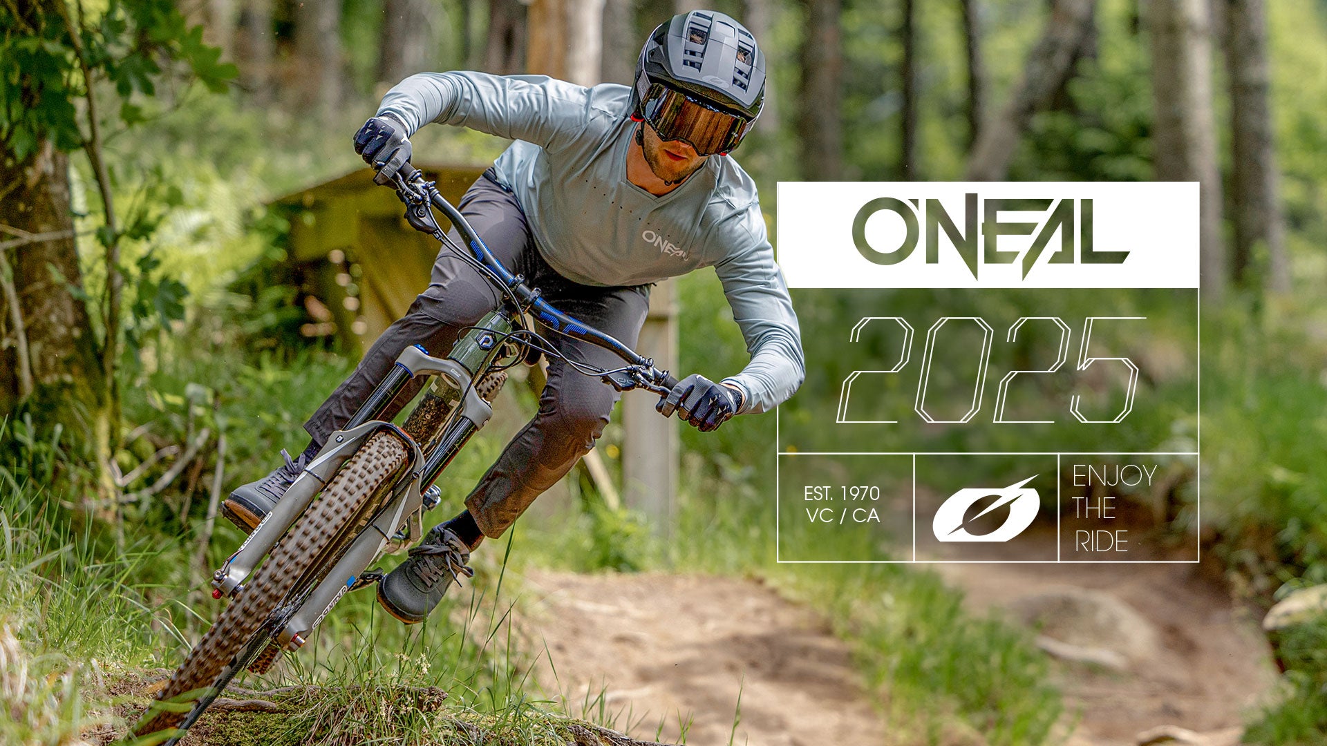 Ein Mountainbiker in voller Montur fährt schnell auf einem Waldweg. Das Bild zeigt den Text „O’NEAL 2025“, „Enjoy the Ride“ und Firmendetails auf der rechten Seite. Bäume und Grün umgeben das Geschehen.