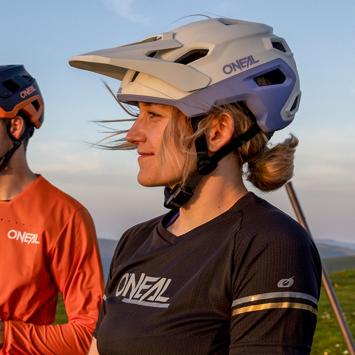 Eine Frau mit weißem O'Neal-Fahrradhelm und schwarzem O'Neal-Trikot steht im Freien und blickt geradeaus. Ein Mann in orangefarbenem O'Neal-Trikot und Helm steht neben ihr. Im Hintergrund sind ein klarer Himmel und eine grüne Landschaft zu sehen.