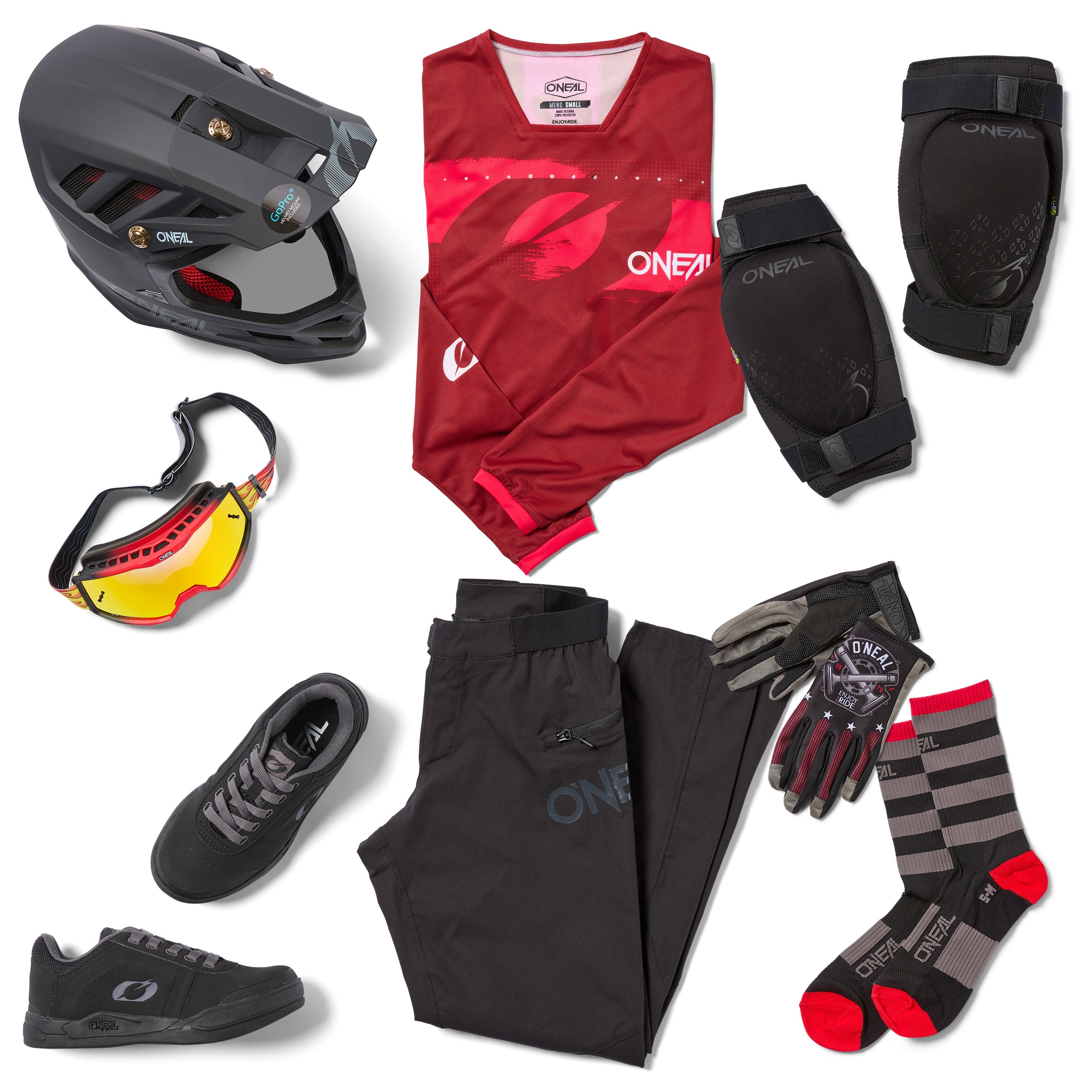 Eine komplette Mountainbike-Ausrüstung liegt bereit, darunter ein Helm, ein rotes Langarmtrikot, eine schwarze Hose, Handschuhe, eine Schutzbrille, Knieschützer, schwarze Schuhe und gestreifte Socken.