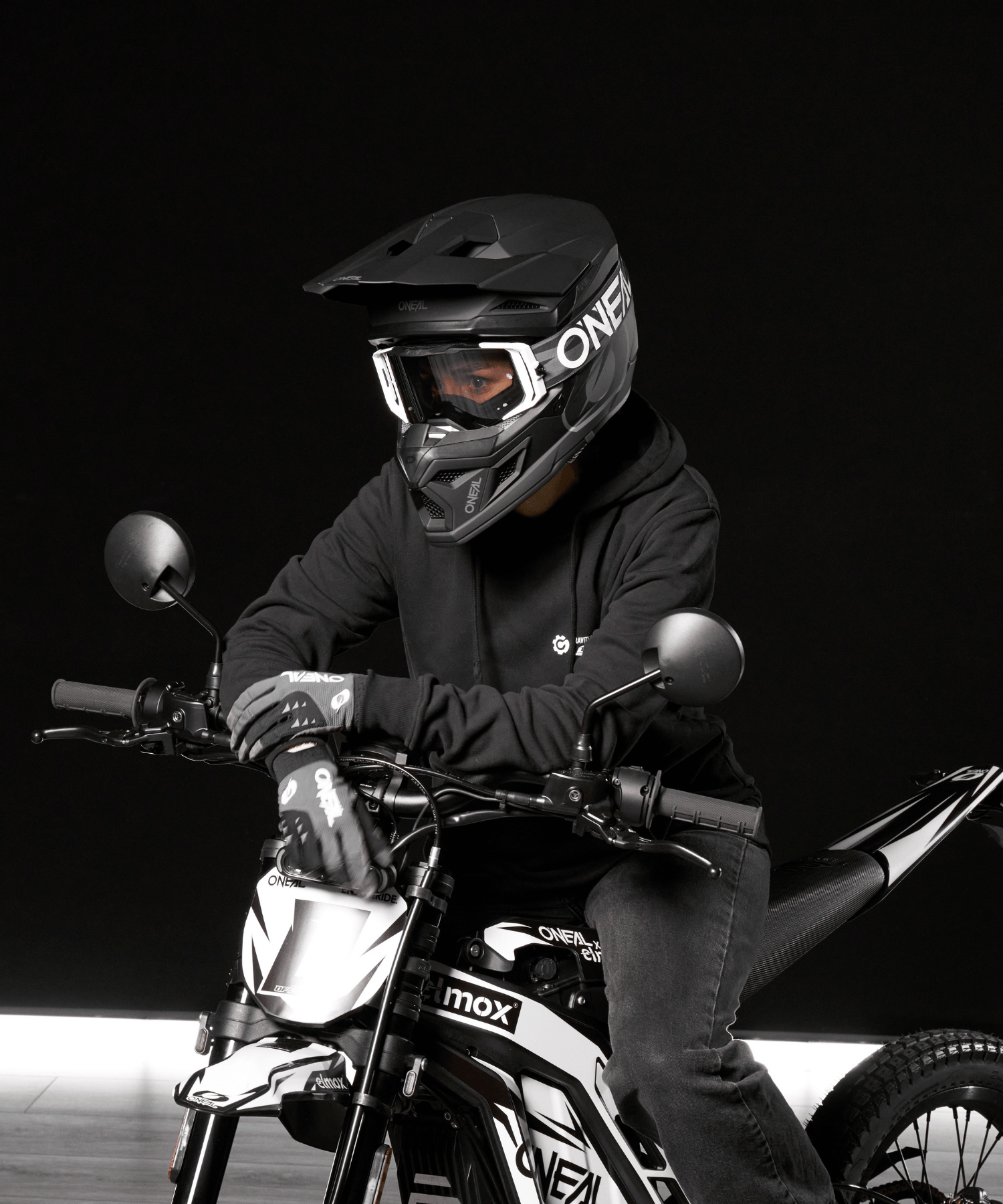 Eine Person mit schwarzem Helm, Schutzbrille, Handschuhen und Kapuzenpullover sitzt auf einem schwarz-weißen Geländemotorrad vor einem dunklen Hintergrund. Das Gesicht des Fahrers ist nicht zu sehen.