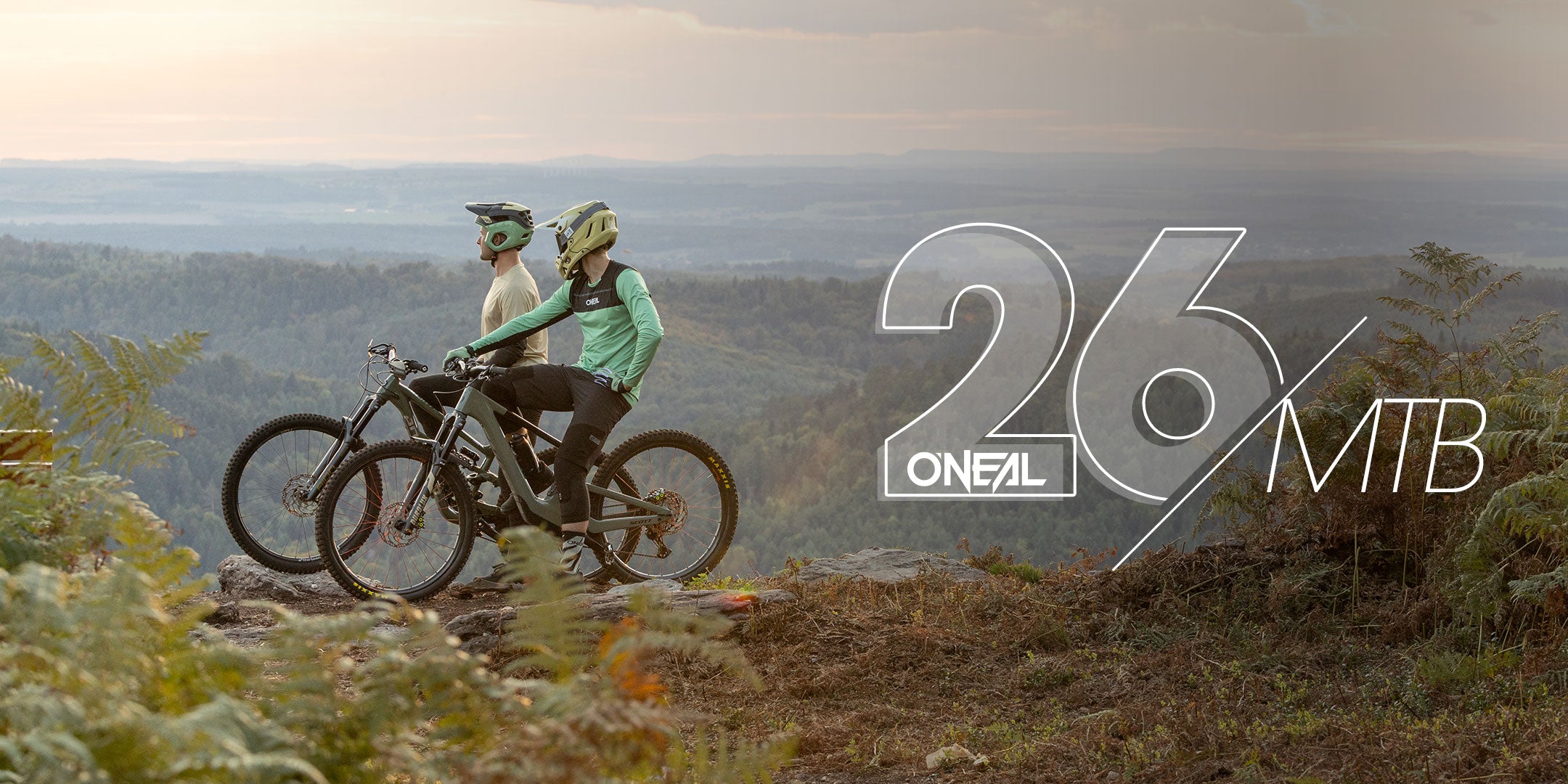 Zwei Personen in Radfahrerkleidung mit Helmen stehen neben ihren Mountainbikes auf einem Felsvorsprung und blicken auf eine malerische Landschaft mit Hügeln und Wäldern bei Sonnenuntergang. Der Text auf dem Bild lautet "26 O'NEAL MTB.