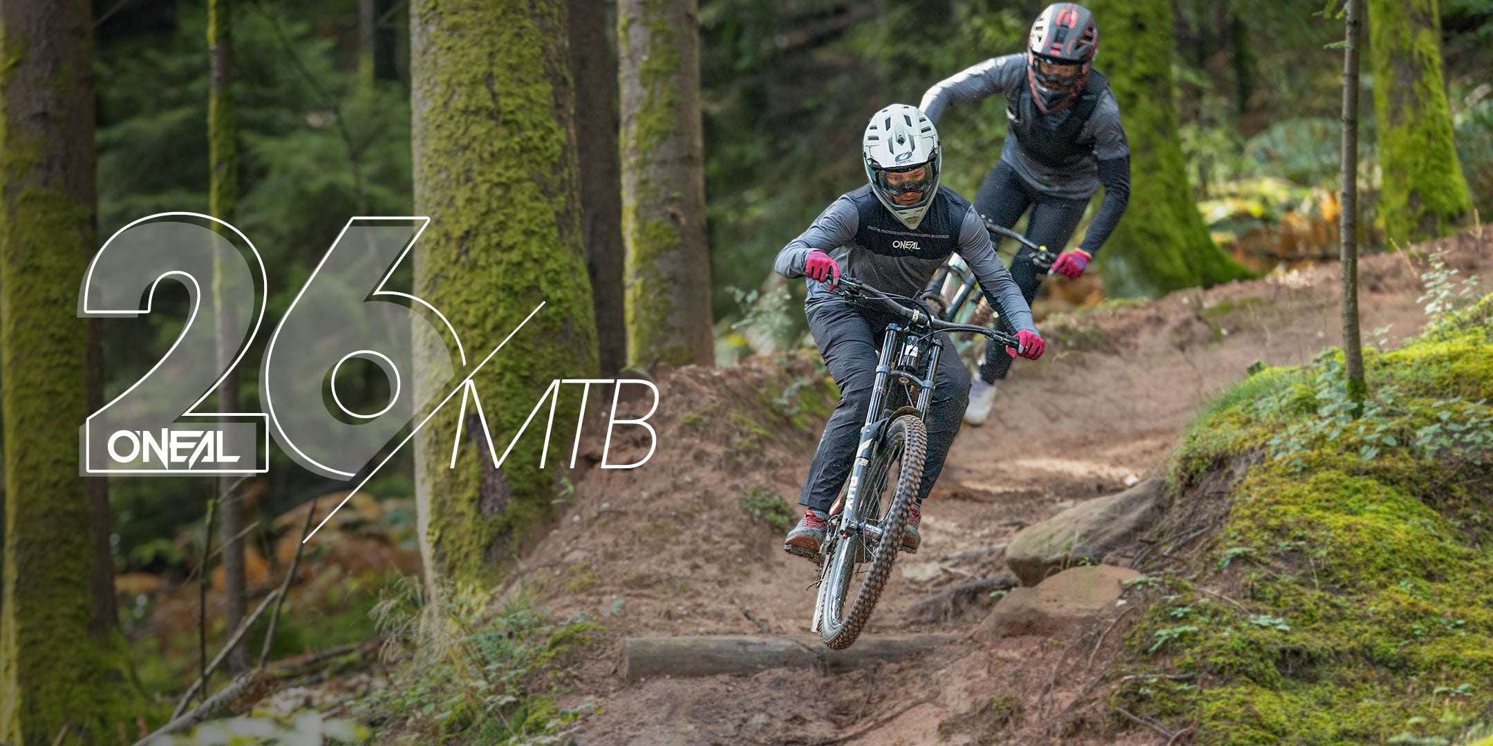 Zwei Mountainbiker mit Helmen und Ausrüstung fahren auf einem unbefestigten Weg durch einen üppigen, grünen Wald. Der überlagerte Text lautet "26 ONEAL MTB". Der Weg ist kurvenreich, und beide Fahrer lehnen sich in eine Kurve.