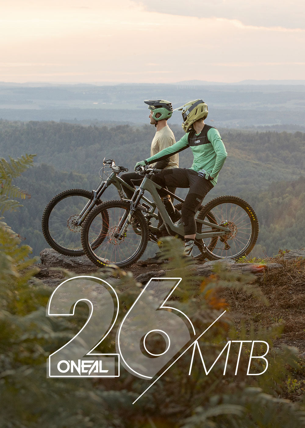 Zwei Mountainbiker mit Helmen und Ausrüstung stehen mit ihren Fahrrädern auf einem felsigen Aussichtspunkt und blicken auf eine malerische, bewaldete Landschaft mit Hügeln in der Ferne. Im Vordergrund steht der Text "O'NEAL 26/MTB".