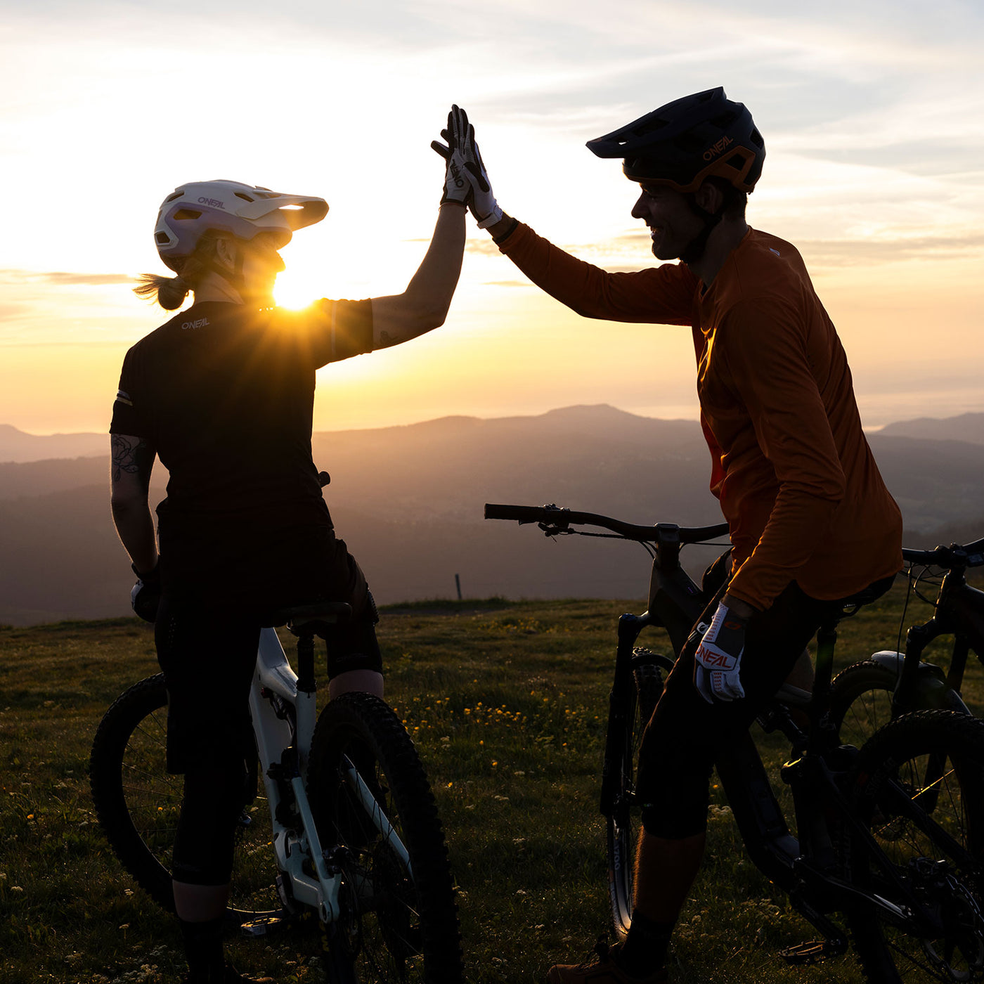 Zwei Radfahrer mit Helmen geben sich bei Sonnenuntergang auf einem Berg ein High Five, neben ihnen sind Fahrräder und im Hintergrund malerische Hügel zu sehen.