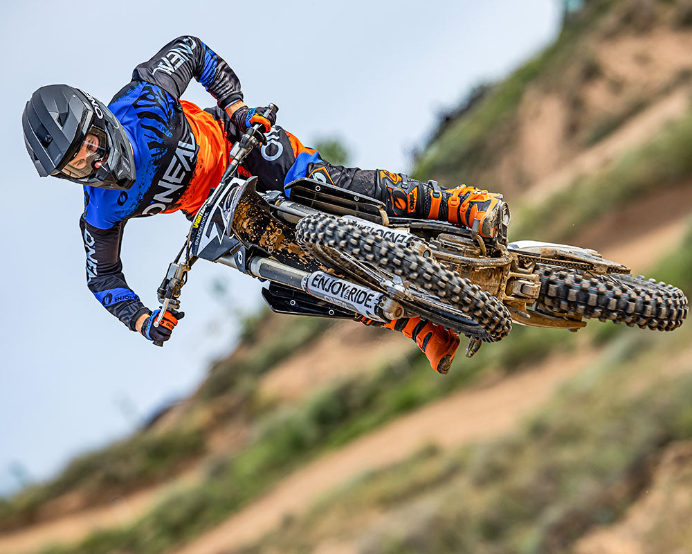 Ein Motocrossfahrer in blau-orangefarbenem Outfit macht einen Hochsprung auf einem Dirtbike. Der Fahrer lehnt sich zur Seite, während das Bike in der Luft geneigt ist. Im Hintergrund sind eine verschwommene Feldwegstrecke und eine Naturlandschaft zu sehen.