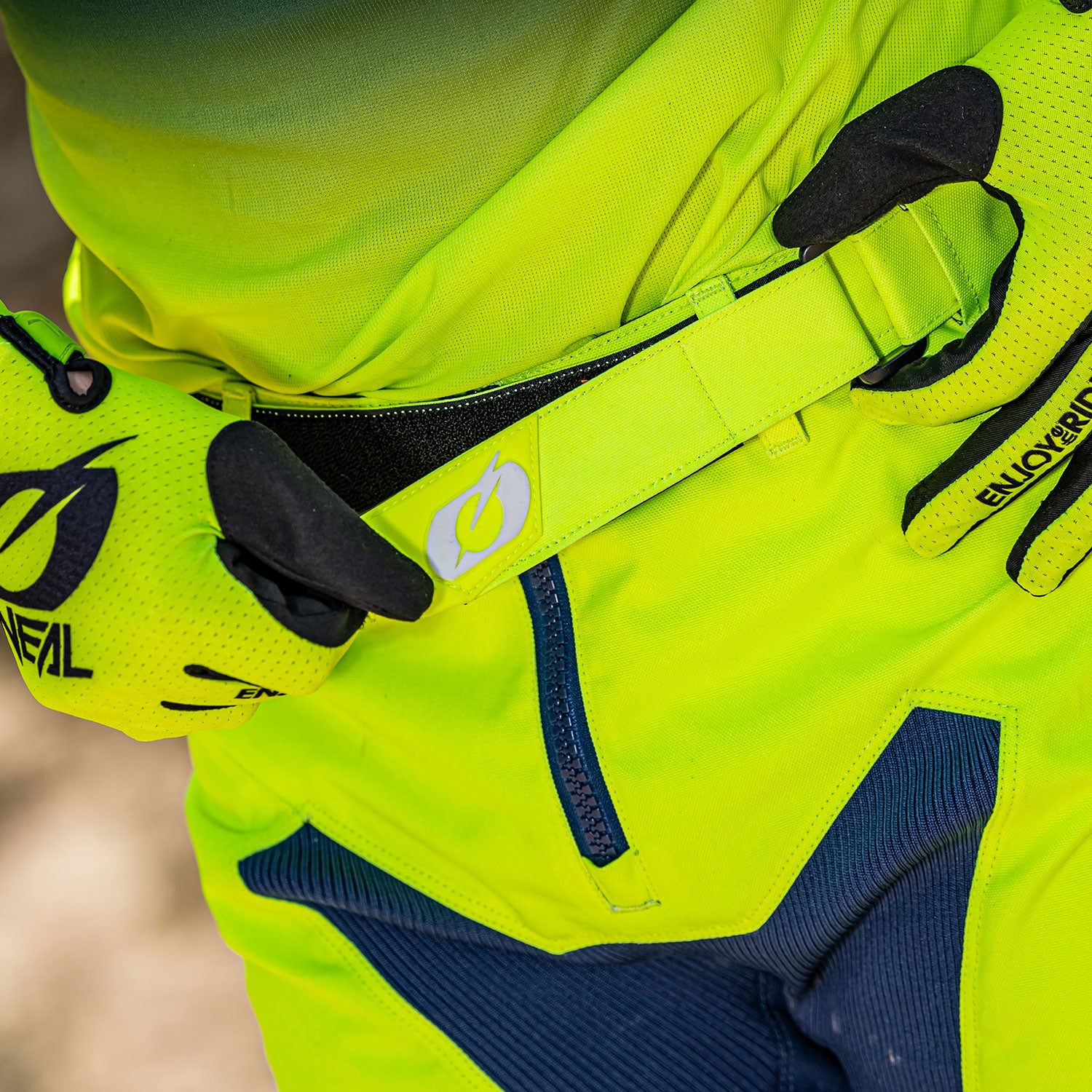 Nahaufnahme einer Person, die leuchtend gelbe Handschuhe und dazu passende Hose und Oberteil trägt und einen Gürtel an ihrer Motocross-Hose zurechtrückt. Der Gürtel und die Handschuhe weisen ein auffälliges „O“-Logo auf. Die Hose hat marineblaue Akzente im Kniebereich.