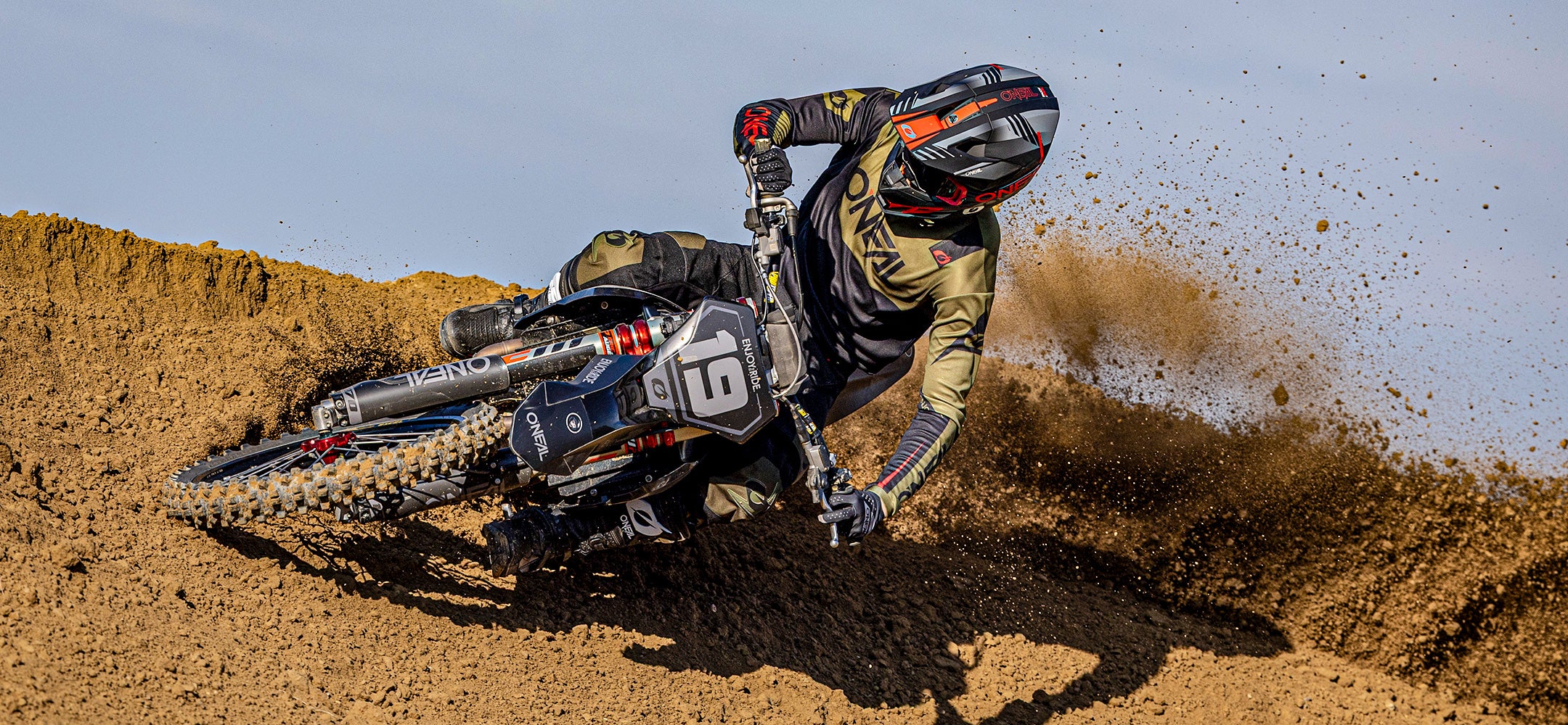Ein Motocross-Fahrer in schwarzer und olivgrüner Kleidung lehnt sich scharf an ein Dirt Bike mit der Nummer 61 und wirbelt bei einem Rennen auf einer Motocross-Strecke unter freiem Himmel eine Menge Schmutz auf.