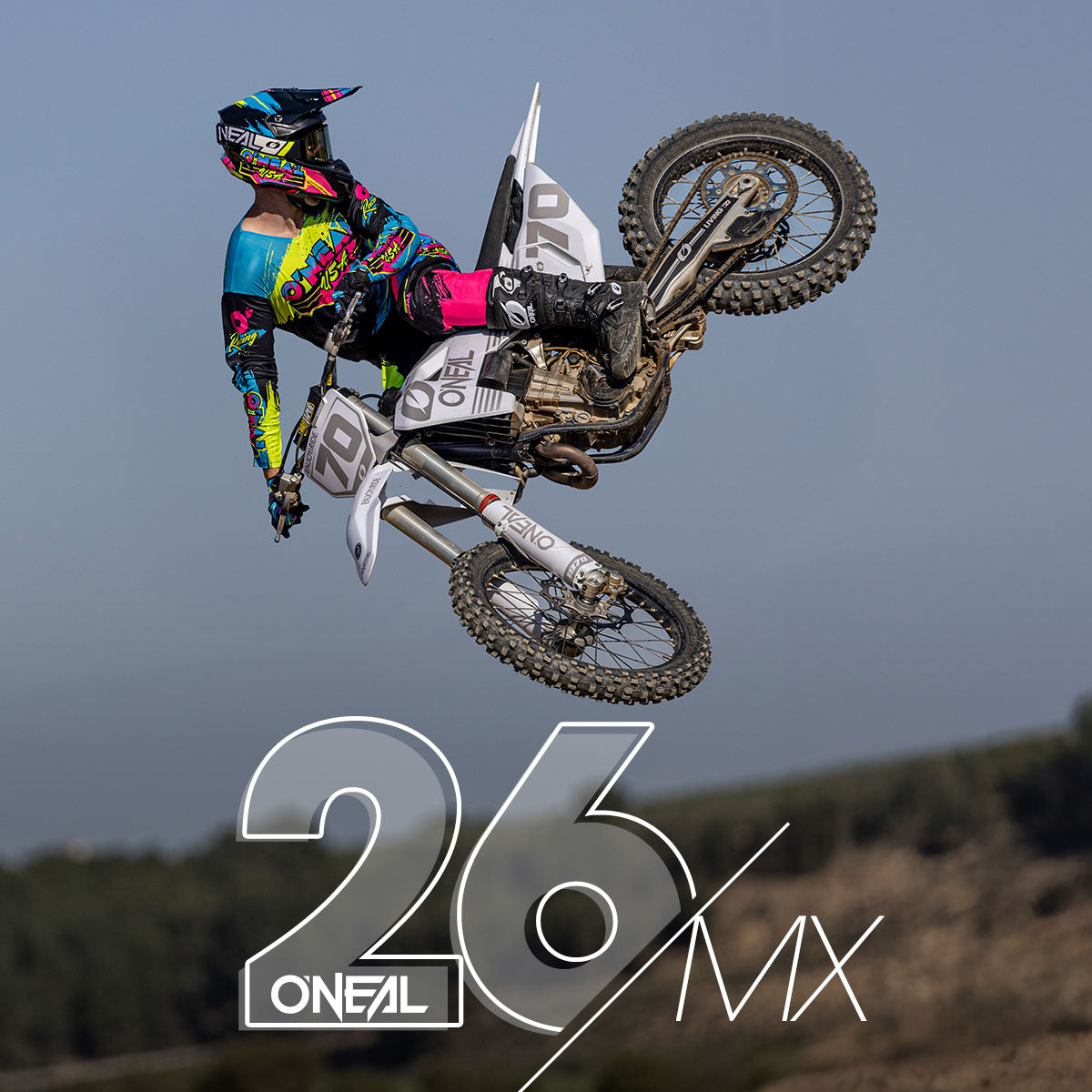 Ein Motocross-Fahrer in farbenfroher Ausrüstung vollführt einen Trick in der Luft auf einem Dirt Bike, seitlich angewinkelt gegen einen klaren Himmel. Die Nummer 26 und der Schriftzug "ONEAL MX" sind prominent im Vordergrund zu sehen.