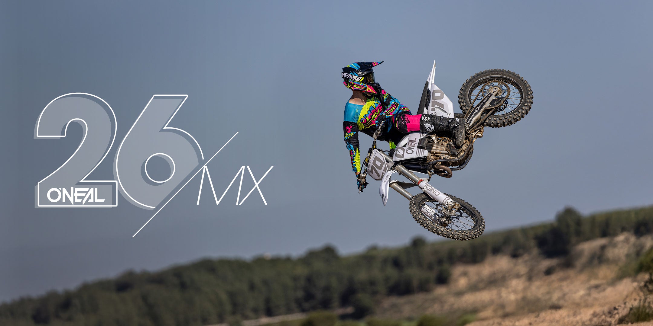 Ein Motocross-Fahrer in farbenfroher Ausrüstung vollführt einen hohen Sprung auf einem Dirt Bike vor einem klaren Himmel, mit dem Text "26 MX ONEAL" auf der linken Seite und einer verschwommenen Landschaft im Hintergrund.