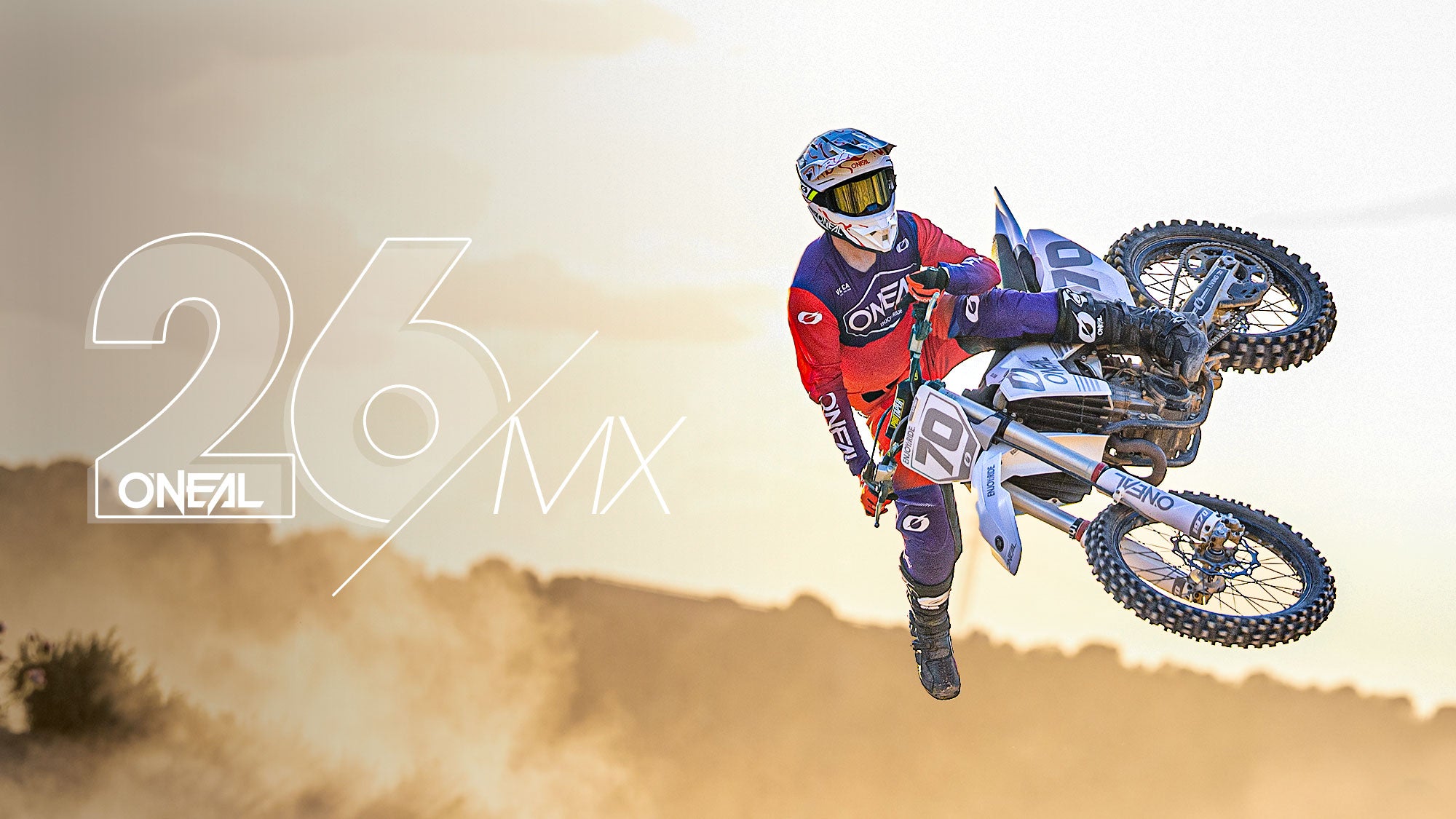 Ein Motocross-Fahrer in roter und blauer Ausrüstung vollführt einen Stunt in der Luft auf einem Dirt Bike, mit Staub im Hintergrund und dem Text "26 O'NEAL MX" auf der linken Seite überlagert.