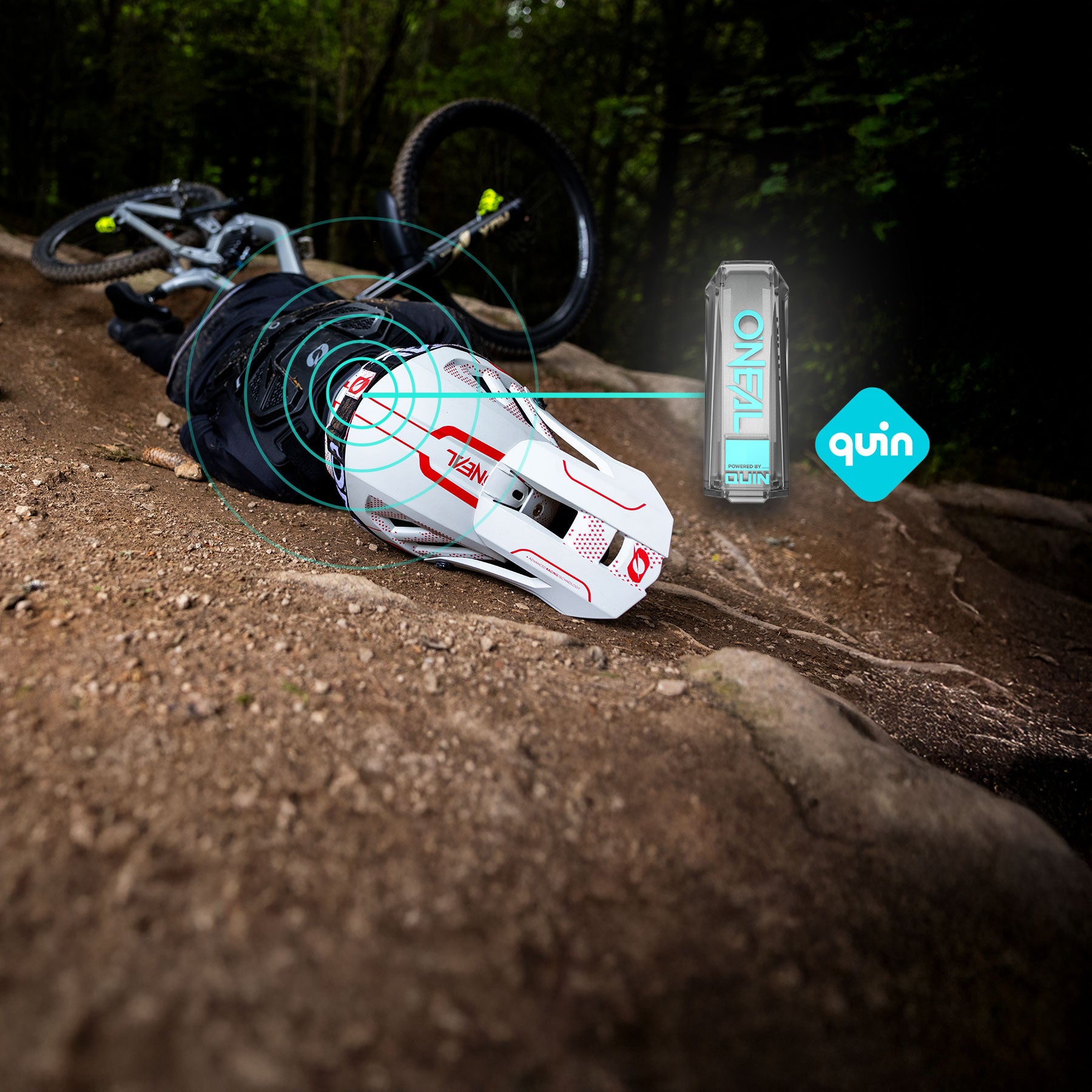 Ein Mountainbiker liegt nach einem Sturz auf einem Feldweg. Sein weißer Helm sendet blaue Signallinien zu einem nahegelegenen Sensorgerät mit der Aufschrift „ONEAL“ und „quin“, was auf einen intelligenten Helm mit Unfallerkennungstechnologie schließen lässt.