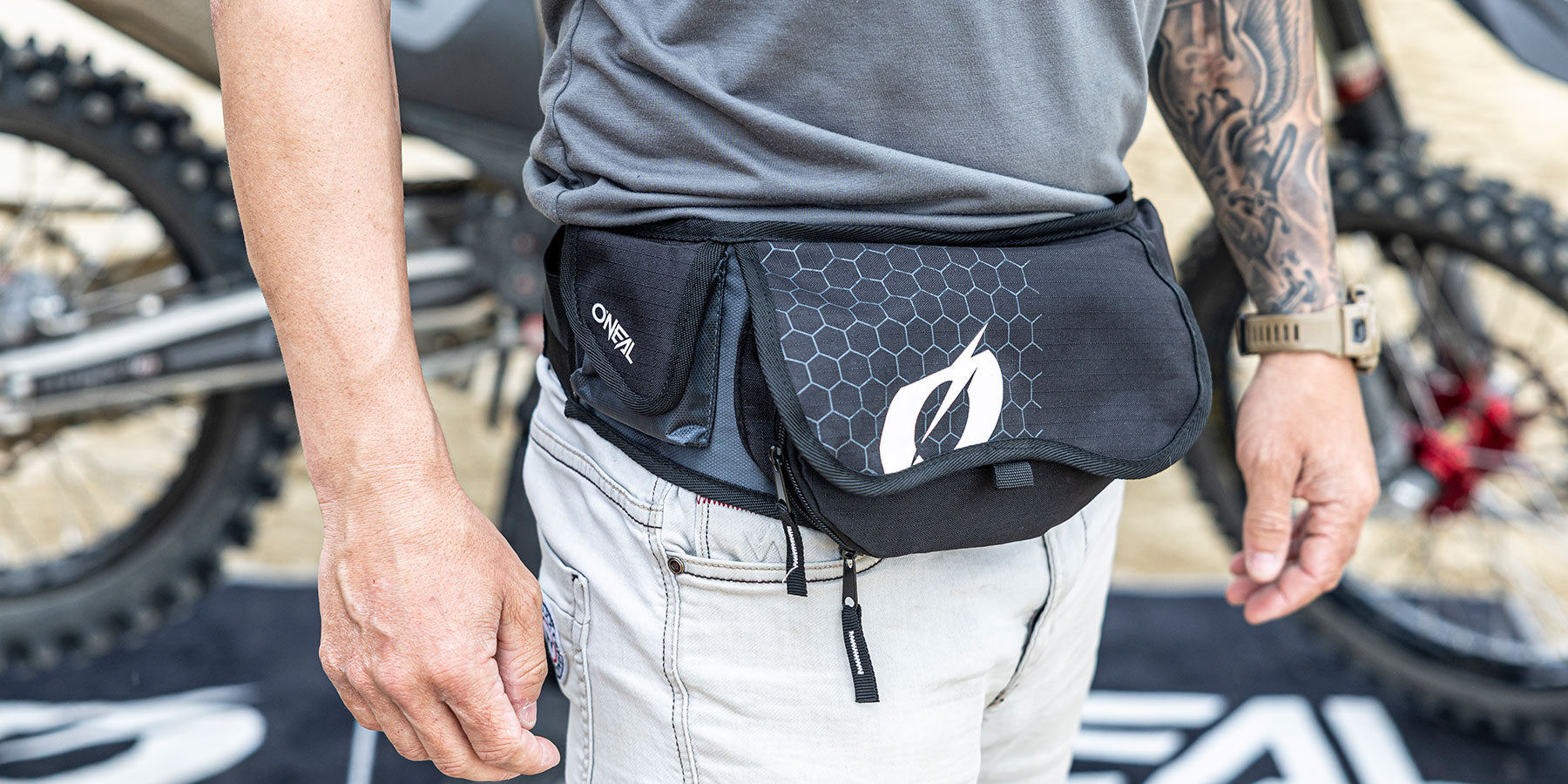 Eine Person, die eine schwarze O'Neal-Gürteltasche mit einem Sechseckmuster trägt, steht vor einem Mountainbike. Die Person ist mit einer hellen Jeans und einem grauen Hemd bekleidet und trägt eine Smartwatch am linken Handgelenk.